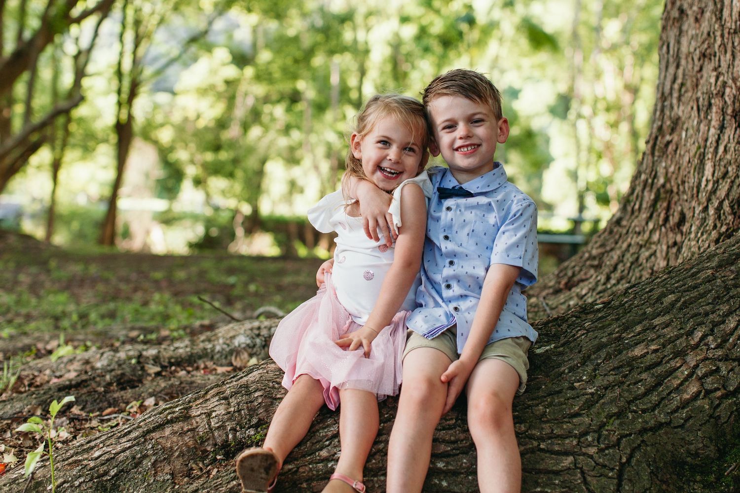 Two happy children sitting together against a large tree in a lush green park, wearing dressy casual outfits.