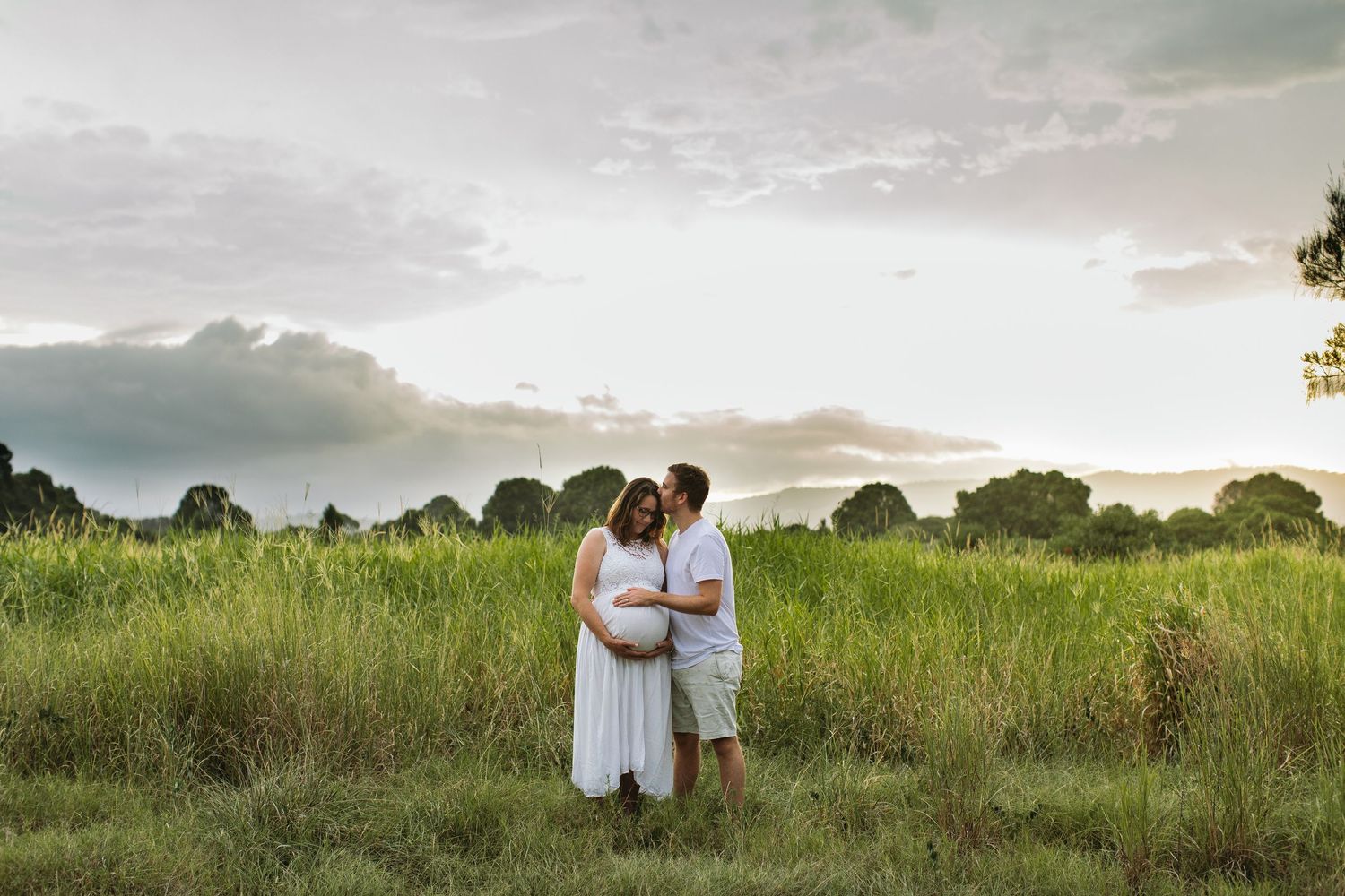 Couple embracing in lush green field at sunset, pregnant woman in white dress, maternity photo session outdoors.