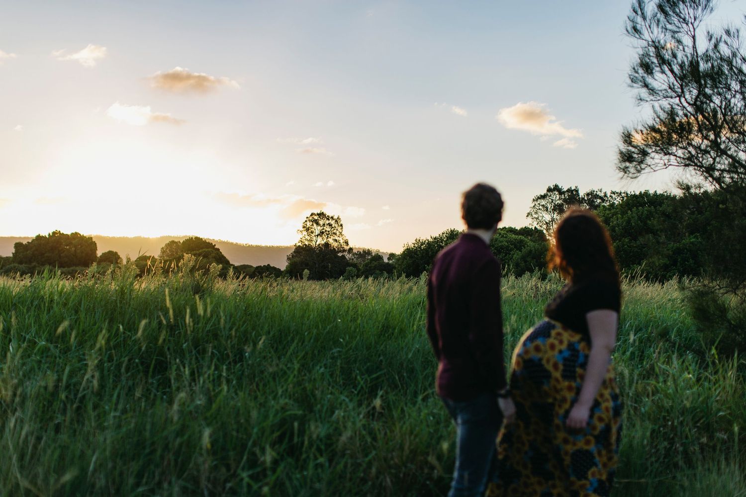 Couple standing in lush green field at golden sunset, surrounded by tall grass and trees.