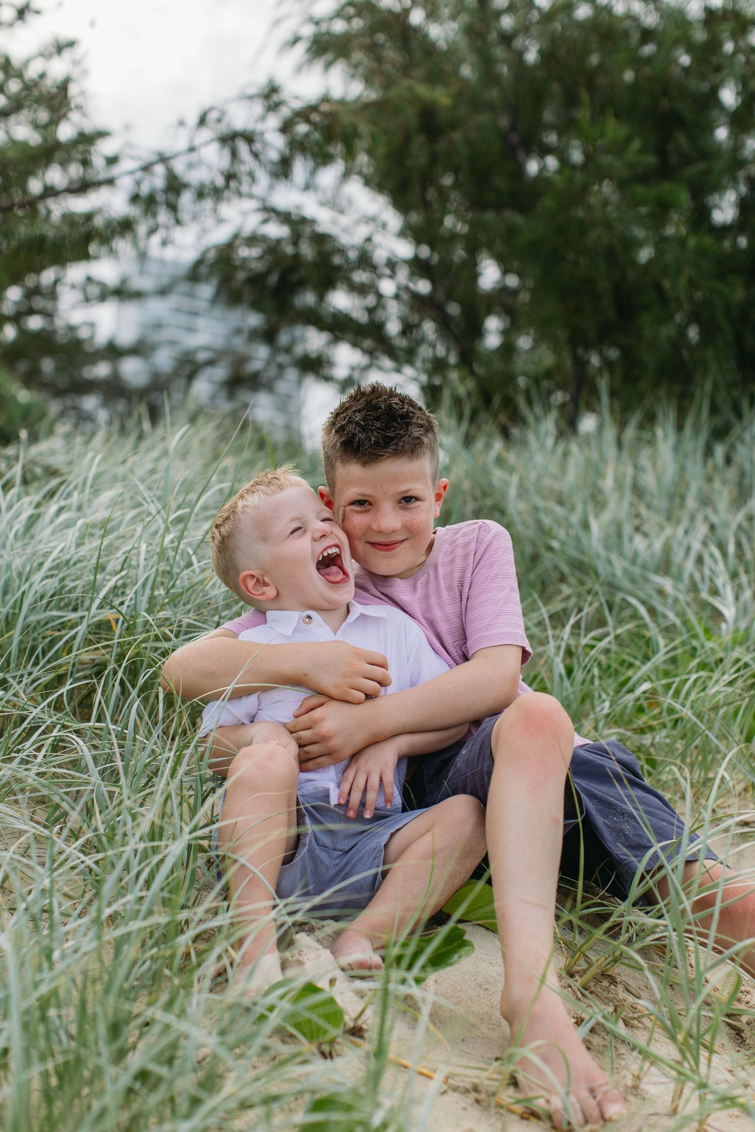 Two brothers laugh and hug while sitting together in tall beach grass on a sandy dune.