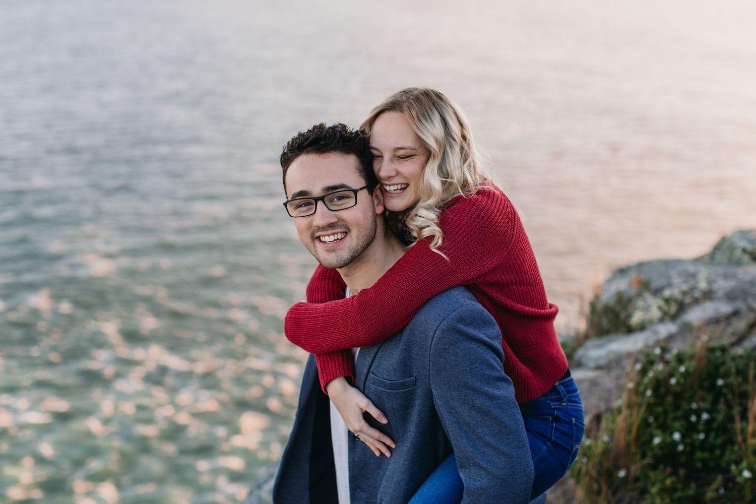 A smiling man with glasses gives a happy blonde woman a piggyback ride near a rocky shoreline.