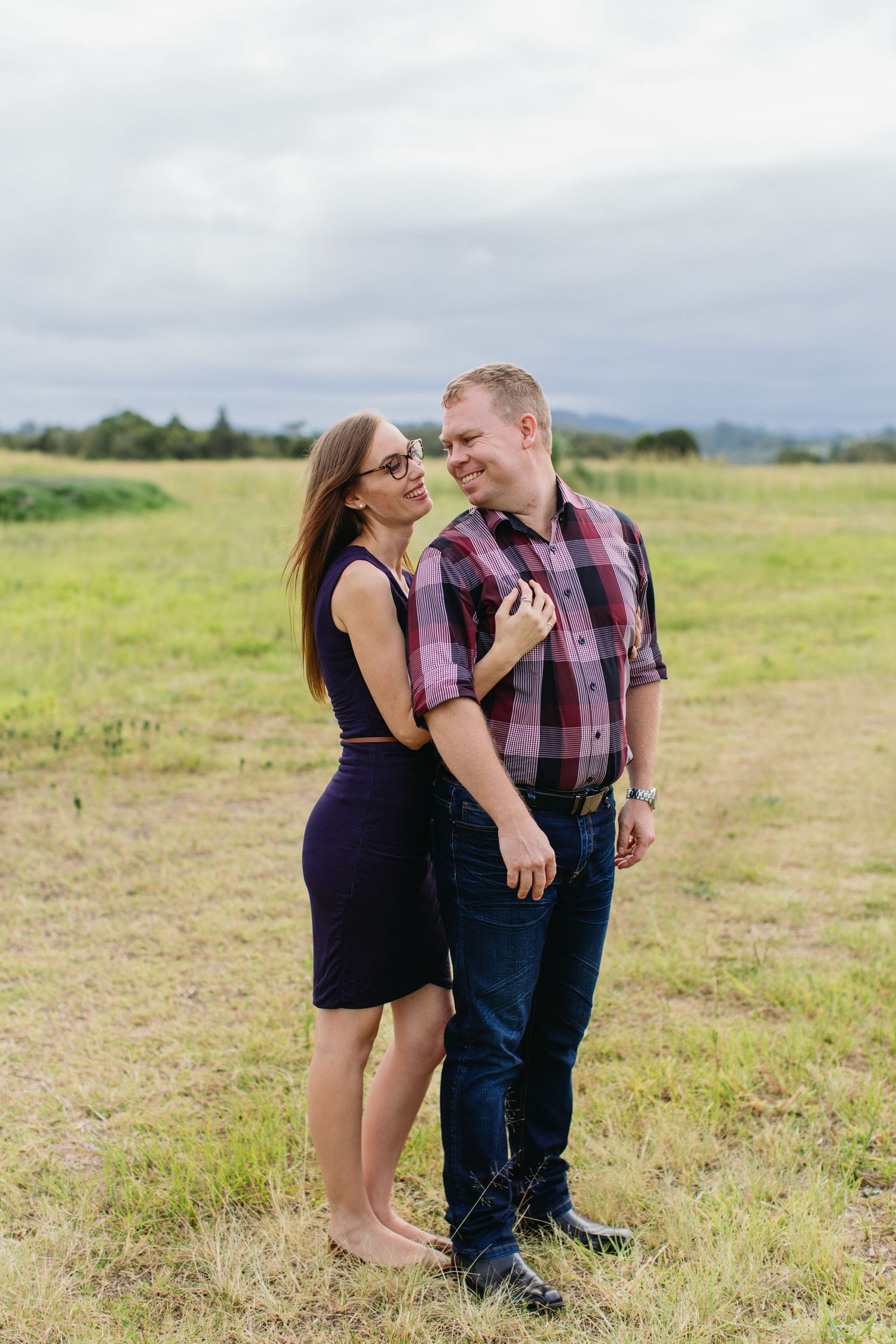 Couple smiling at each other in a green field, woman in purple dress and man in plaid shirt, cloudy sky backdrop.