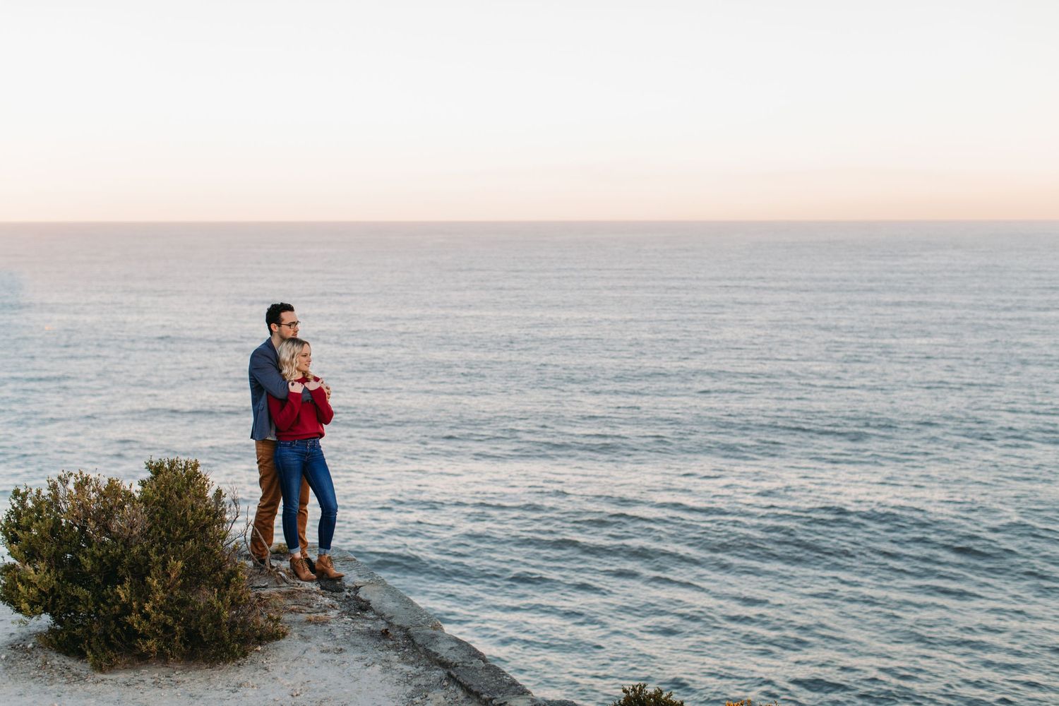 Couple standing on a rocky coastal cliff embracing while enjoying a serene ocean sunset view.