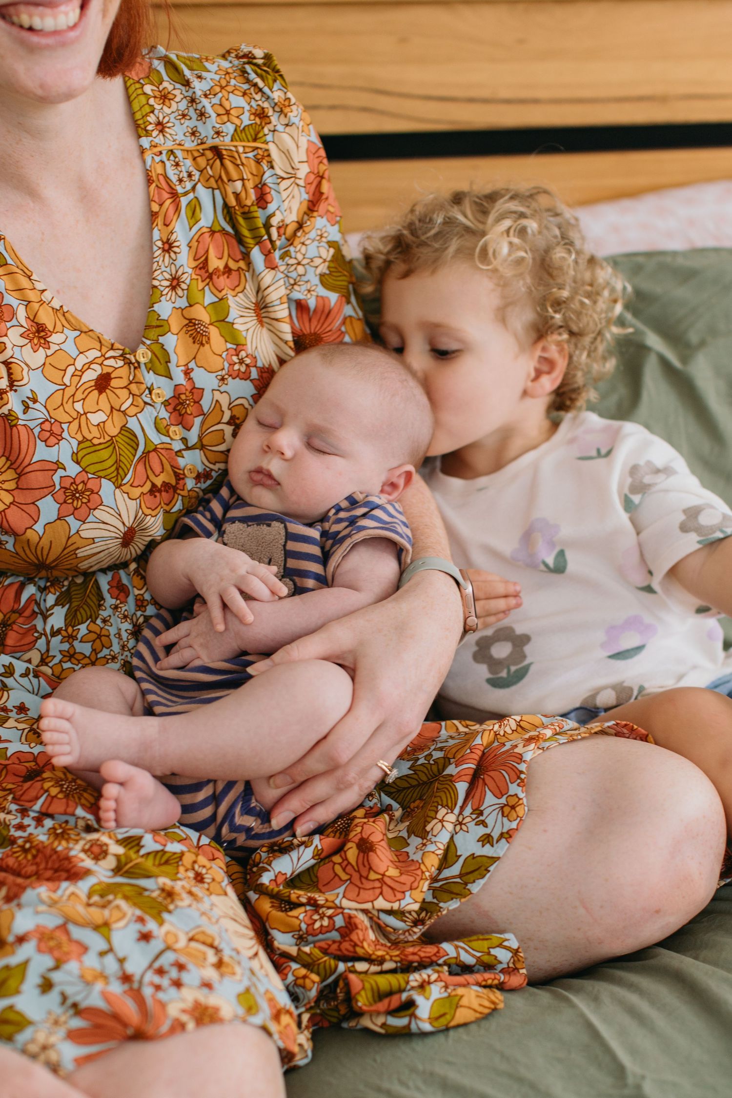 A mother in a floral dress holds a sleeping newborn while a curly-haired toddler gives the baby a gentle kiss.