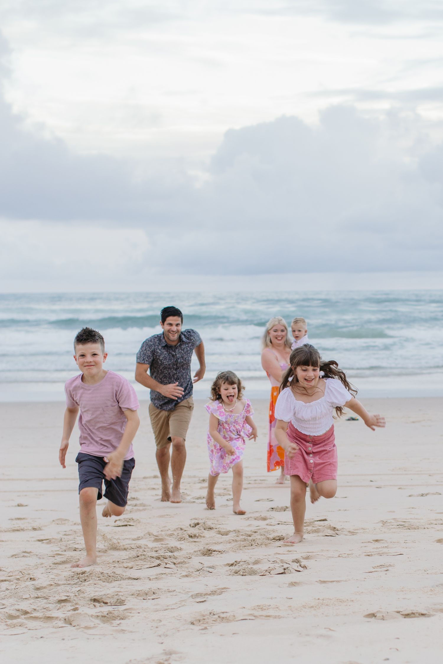 Family of six running and playing on a sandy beach with ocean waves in the background on a cloudy day.