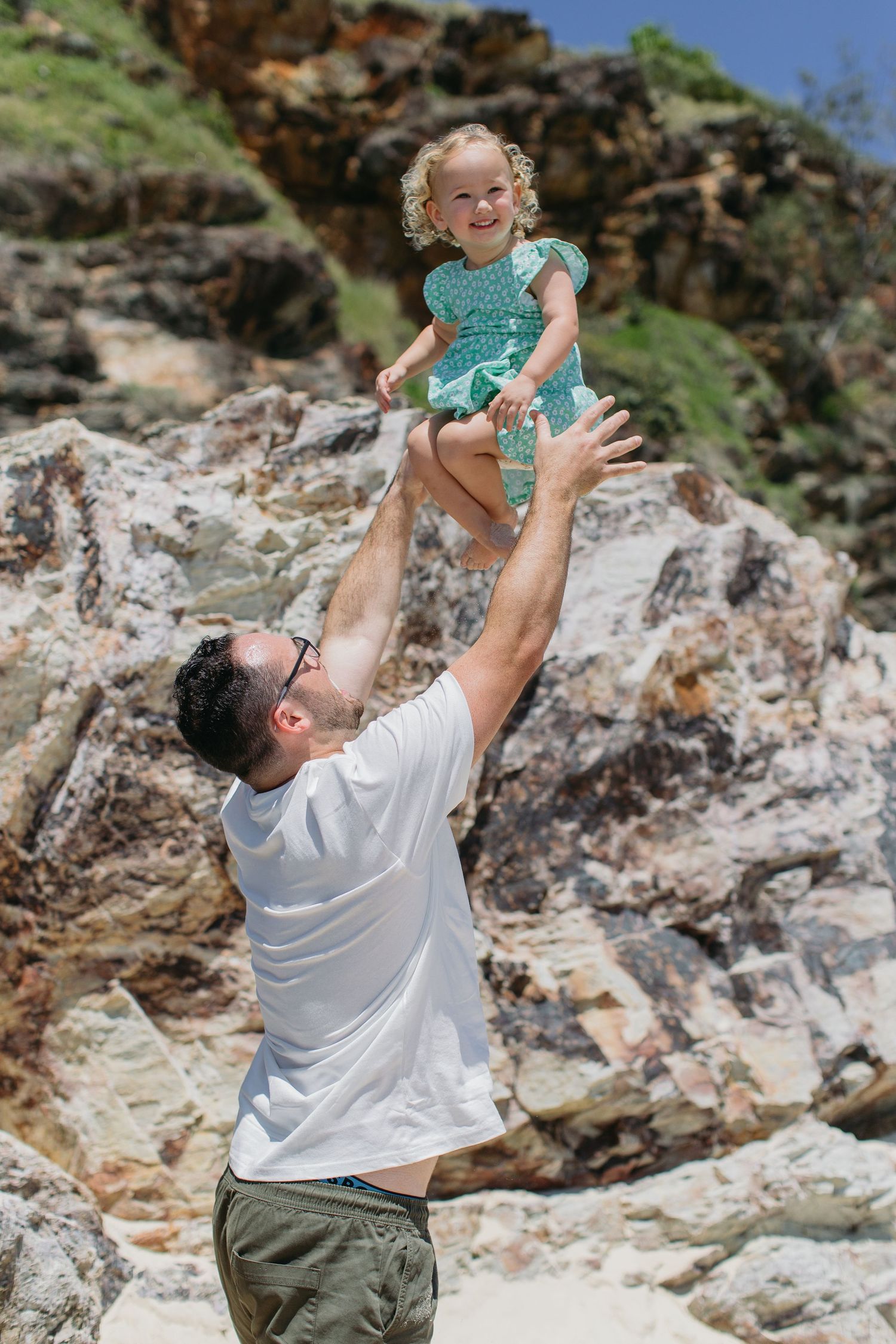 Father tossing laughing toddler girl in teal dress high in the air on a sunny rocky beach.