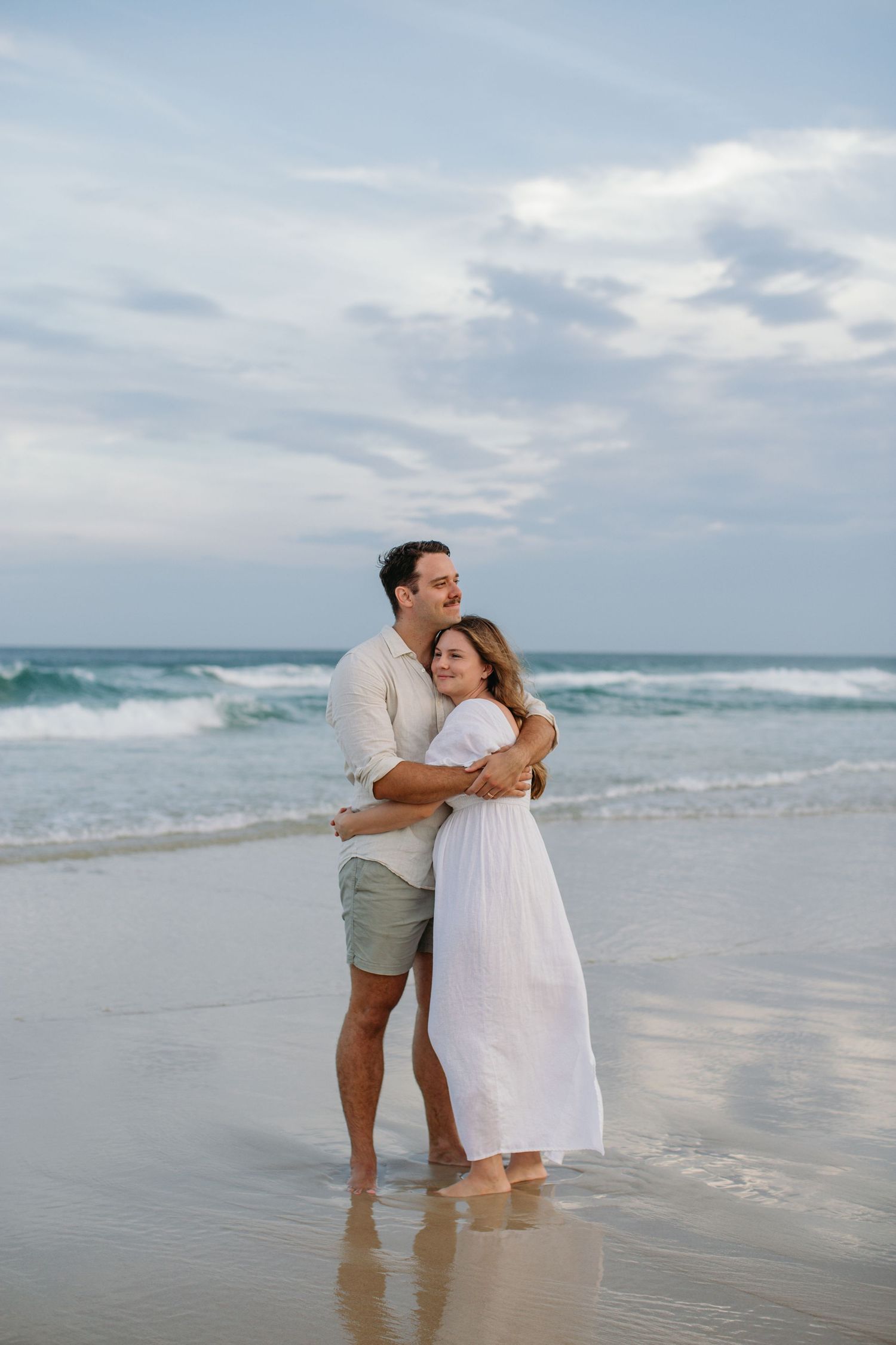 Couple embracing on a sandy beach in white outfits, with ocean waves and cloudy sky in the background.