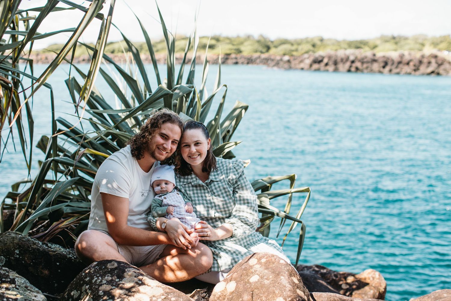 Happy family with baby sitting on rocks by turquoise ocean water near tropical plants on a sunny day.