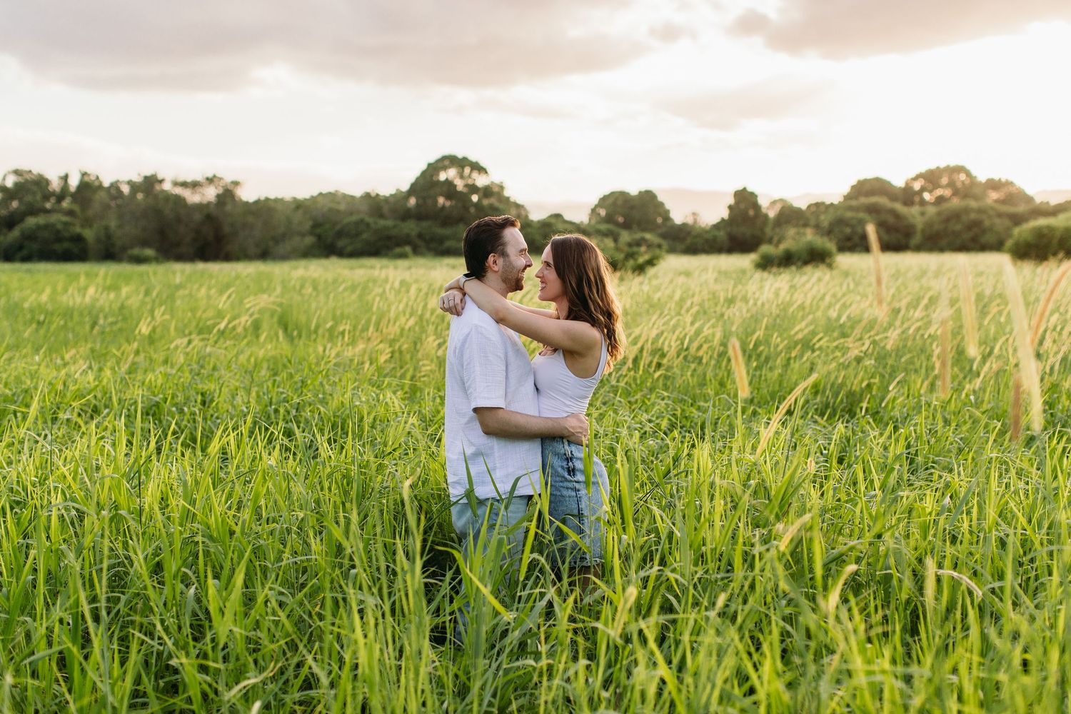 Couple embracing in a lush green field at sunset, sharing a romantic moment surrounded by tall grass and trees.