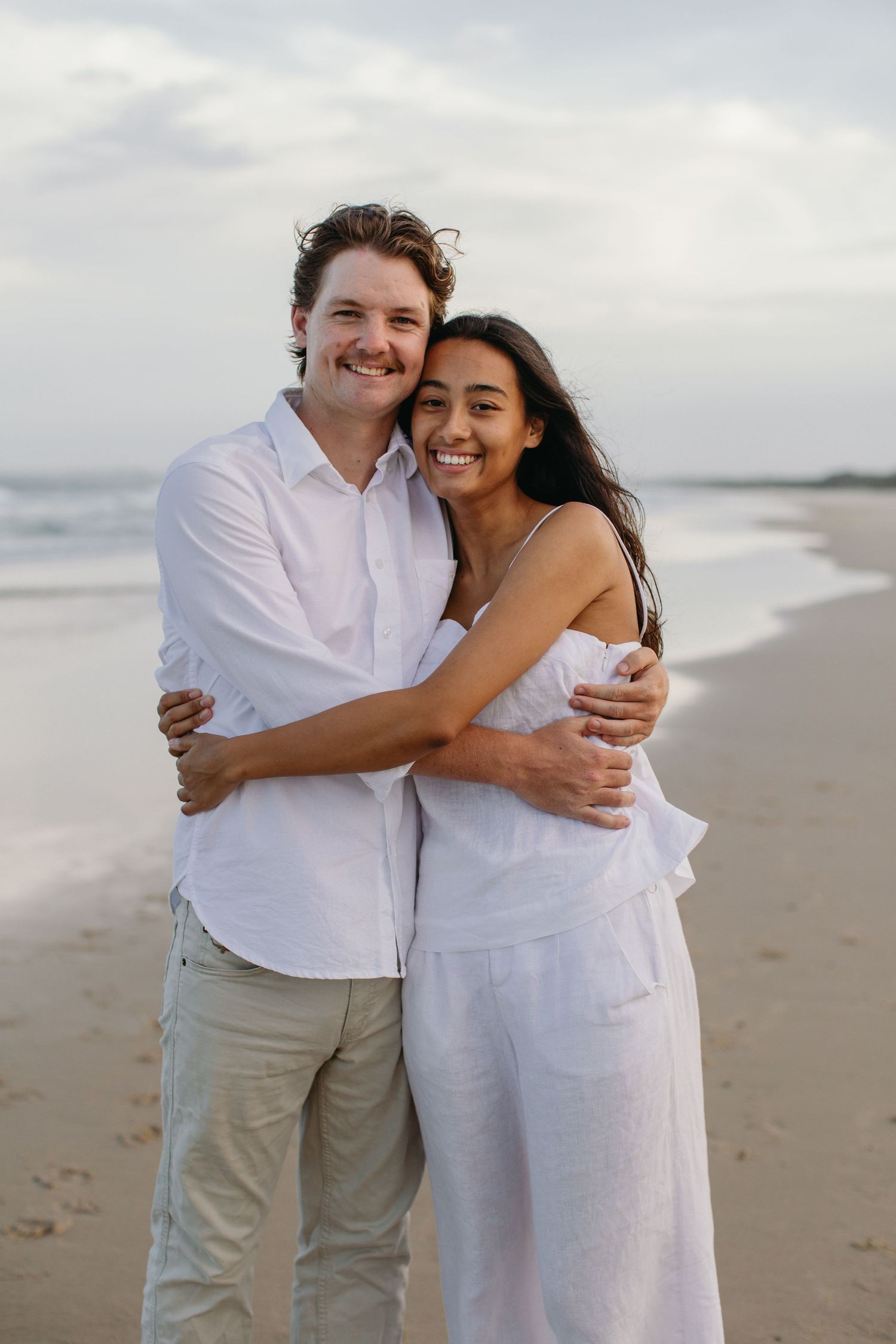 A smiling couple embraces on a sandy beach, both wearing white outfits with ocean waves in the background.
