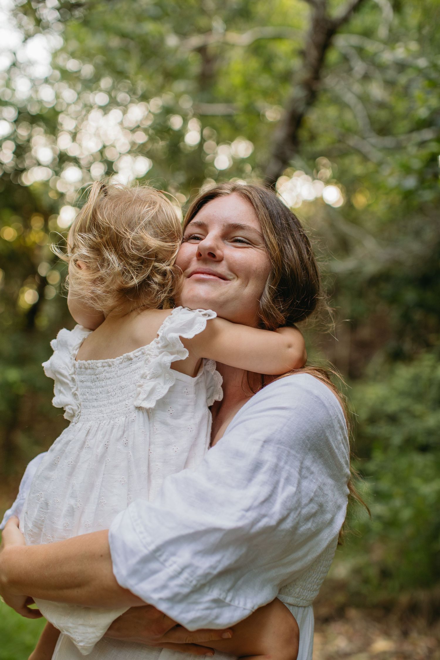 A smiling woman in white linen holds a curly-haired toddler girl in a white dress hugging her cheek outdoors.