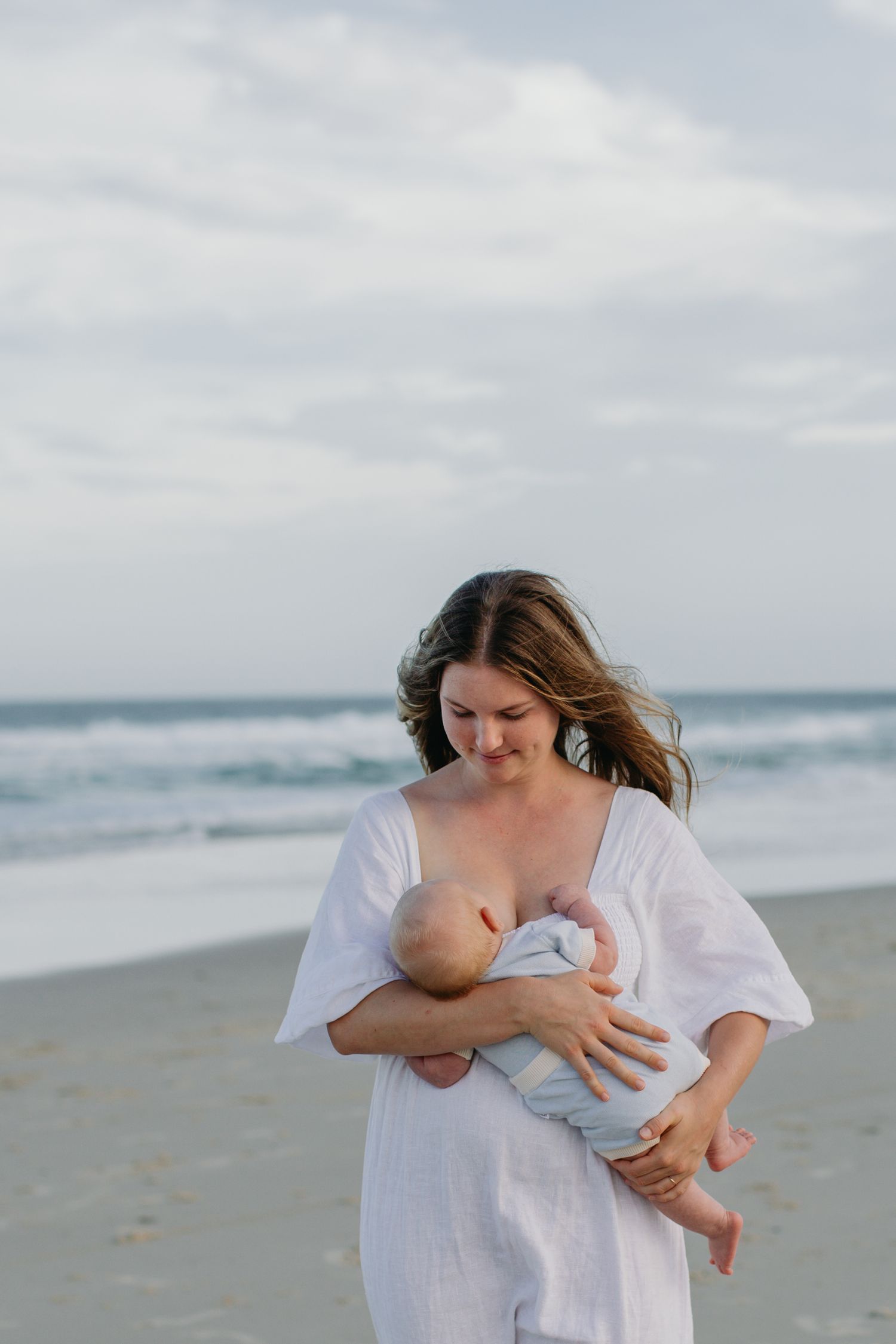 A mother in a white dress tenderly holds her baby on a beach with ocean waves in the background.