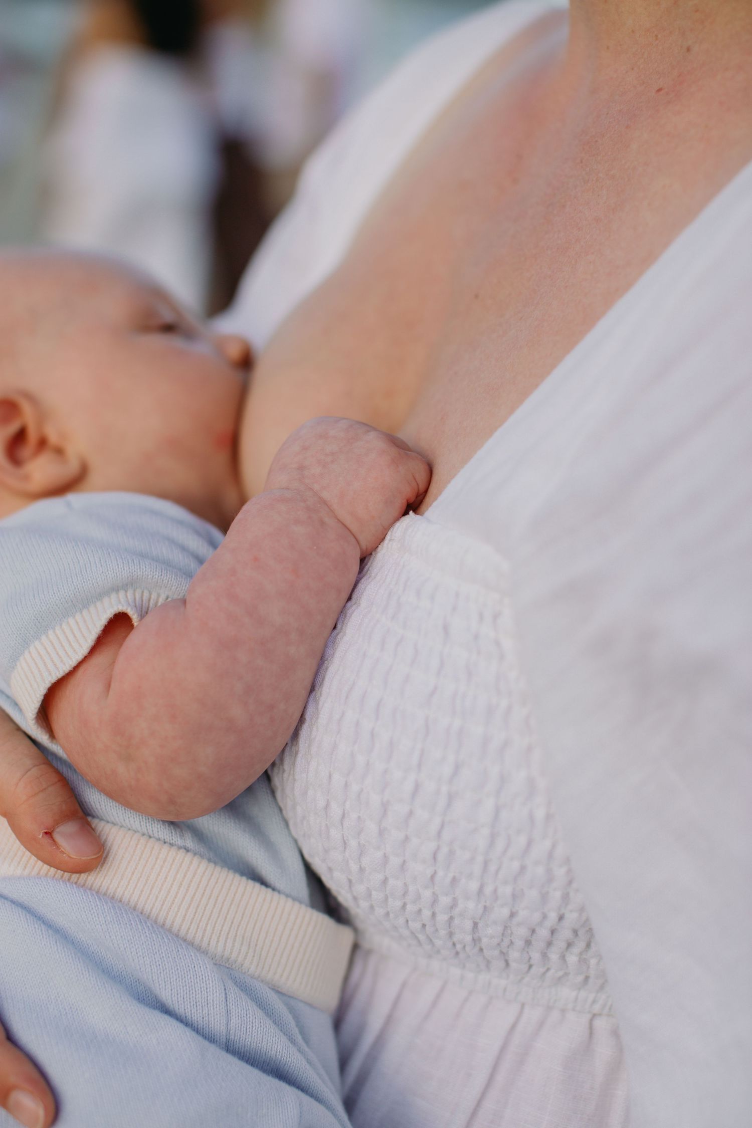 Mother breastfeeding newborn baby in blue outfit, infant's hand resting on mother's white textured top.
