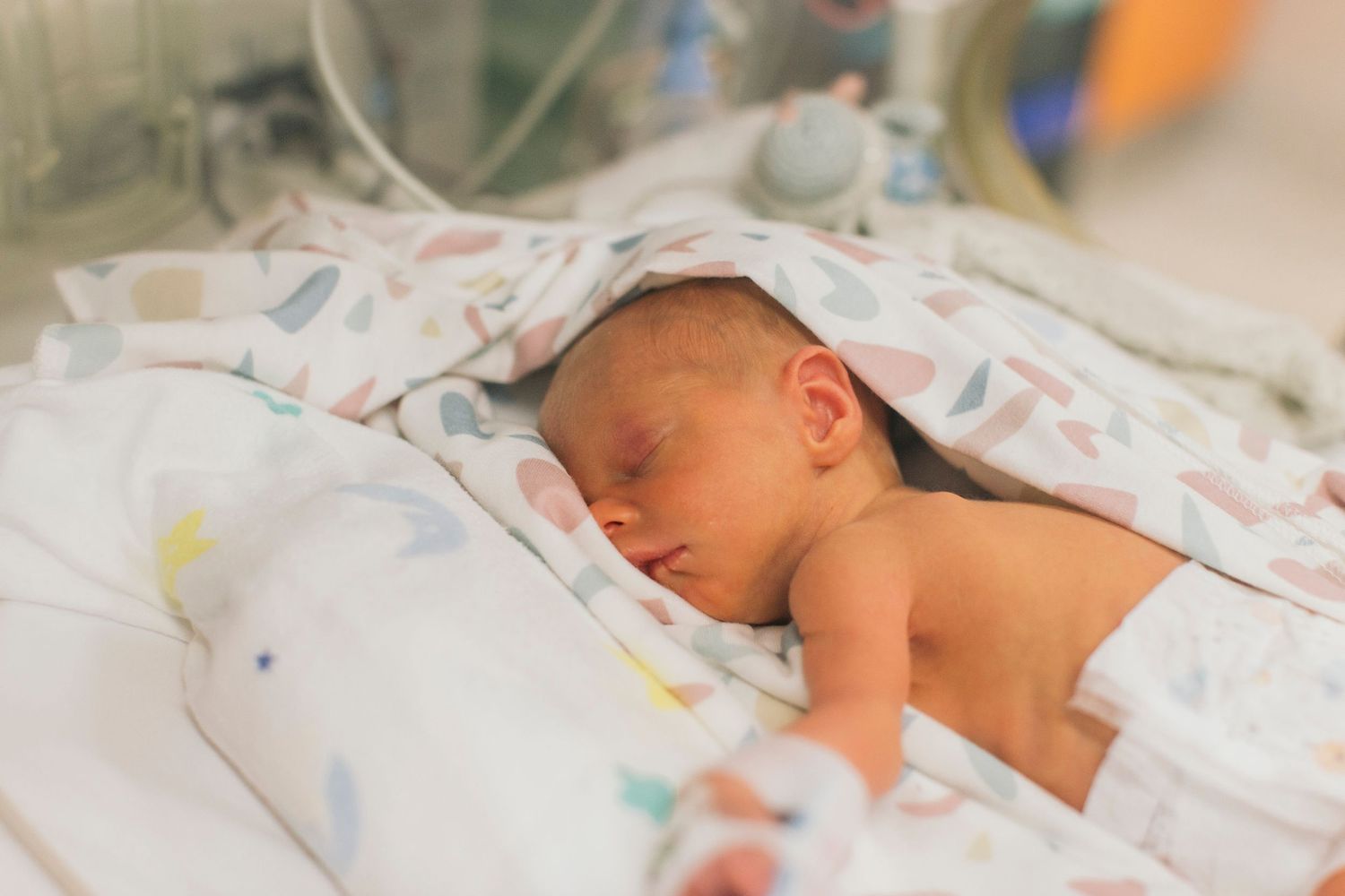 Newborn baby sleeping peacefully in hospital NICU wrapped in colorful blanket under medical care.