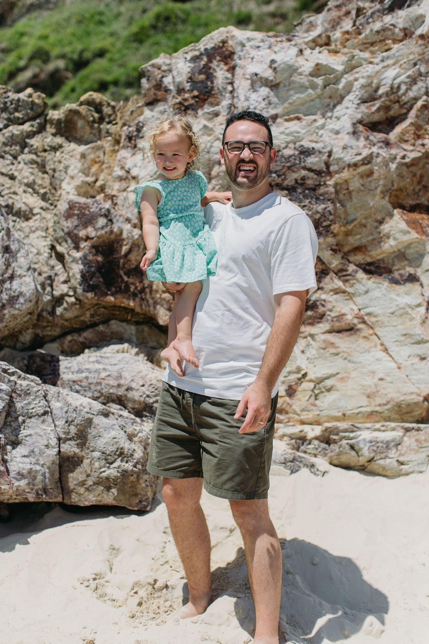 Father holding young daughter in teal dress on sandy beach with rocky backdrop on sunny day.