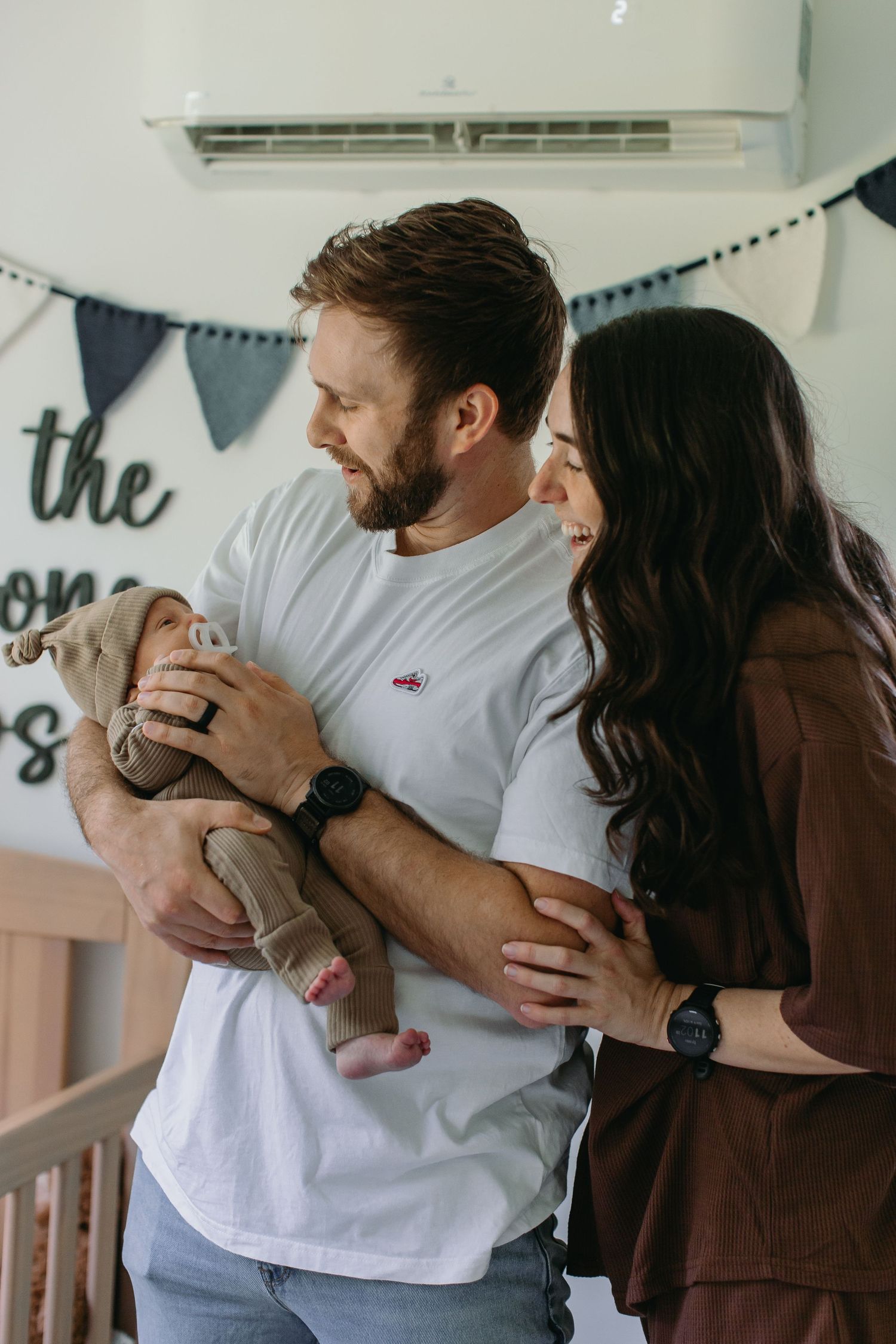 Happy couple in white and brown outfits holding their newborn baby in a decorated nursery room.