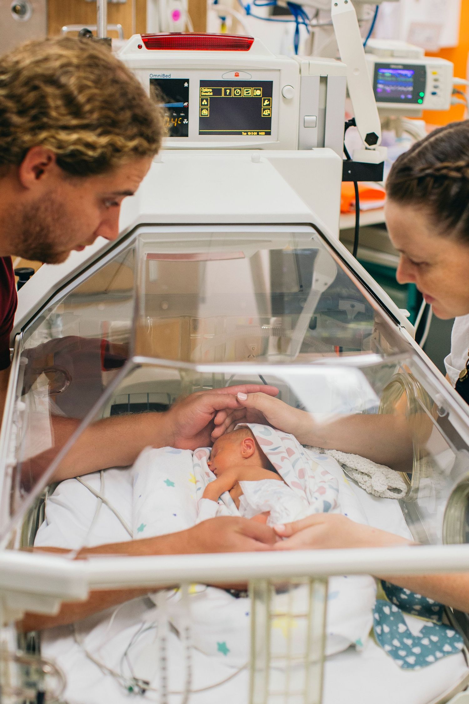 Parents tenderly touch their newborn baby in a hospital incubator in the NICU, surrounded by medical monitoring equipment.