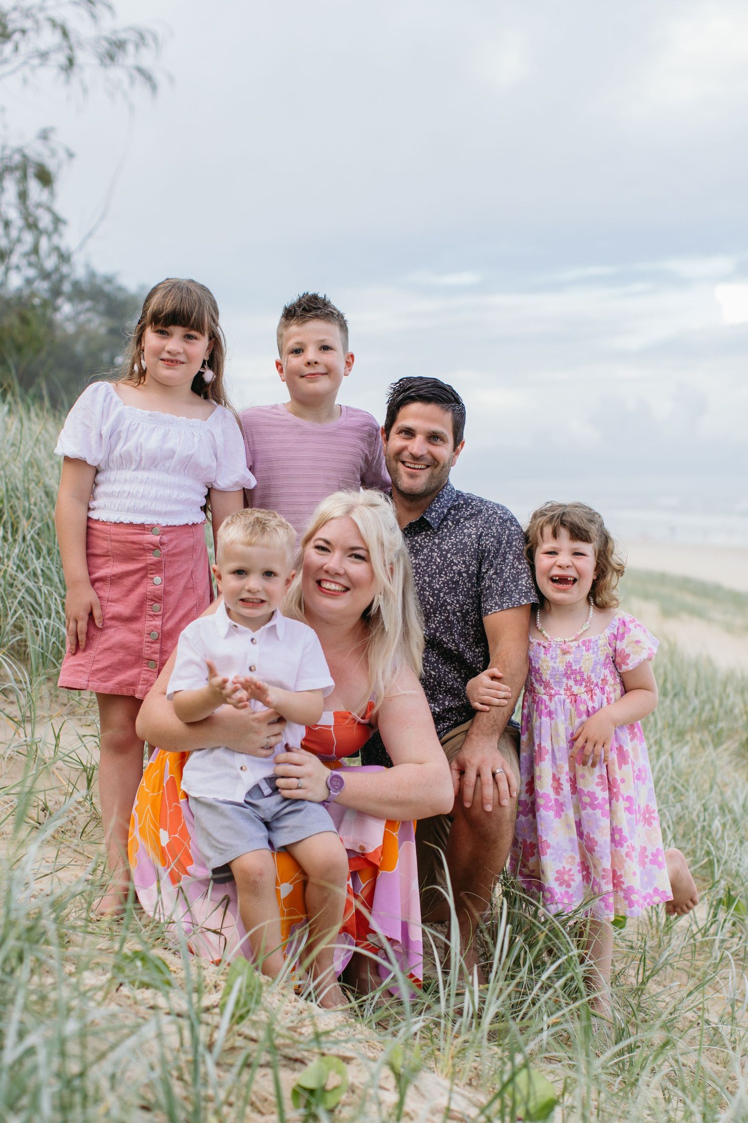 Family of six posing on sandy beach dunes, parents with four young children smiling for a portrait.