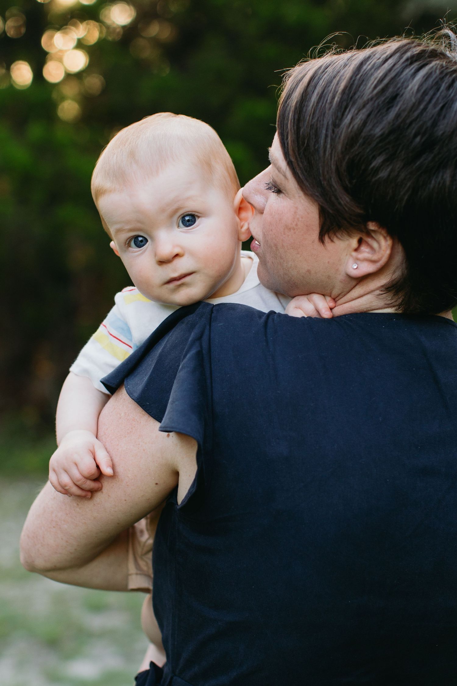 A parent holding a cute blue-eyed baby outdoors, gently kissing the infant's cheek near green trees.