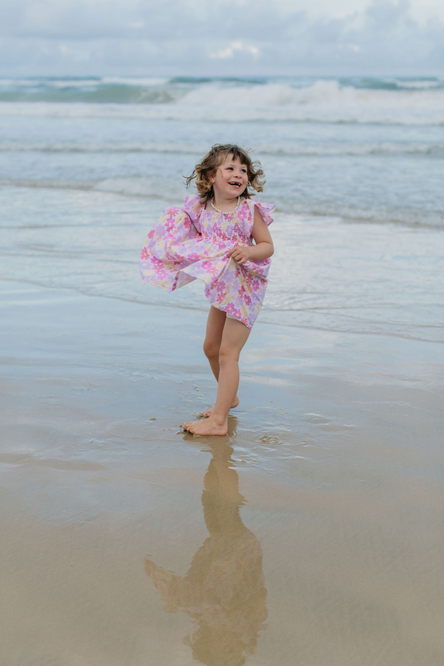 Happy young girl in pink floral dress playing in ocean waves on sandy beach.