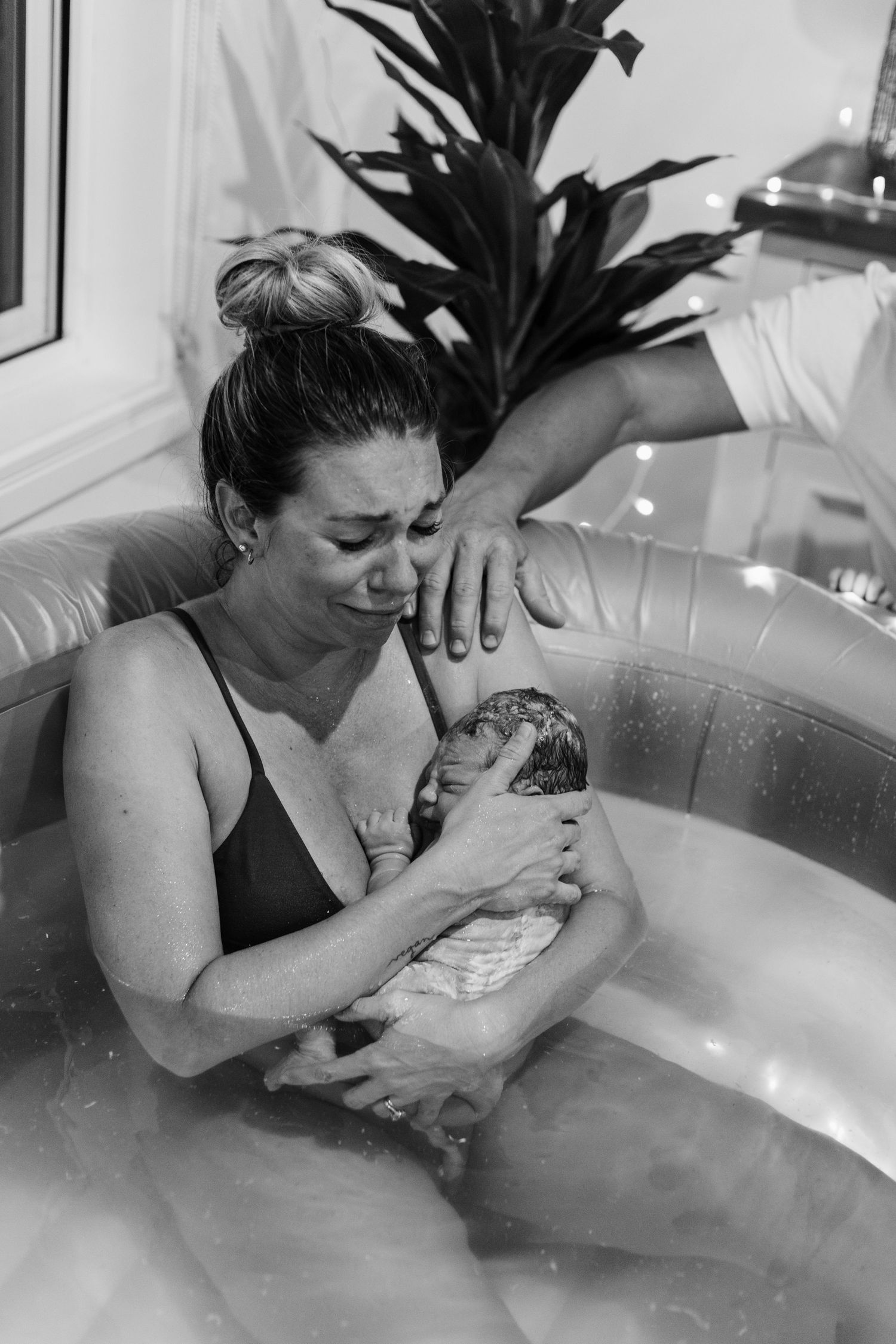 Black and white photo of emotional mother holding newborn in birth pool with supportive hand nearby.