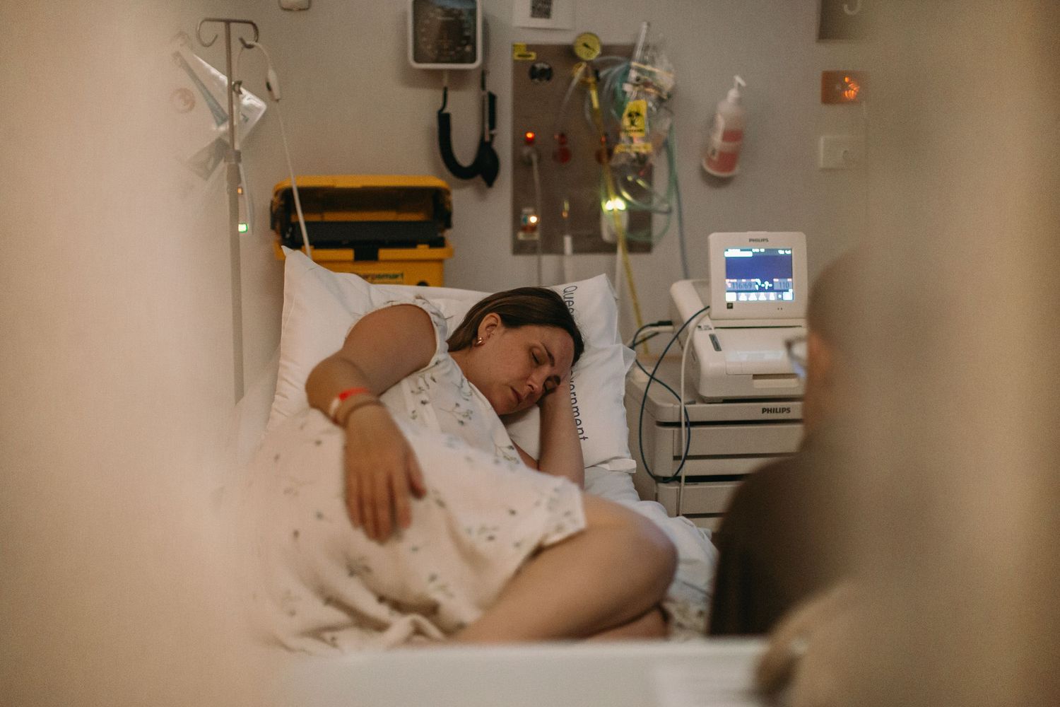 Woman resting in hospital bed surrounded by medical equipment and monitors in a clinical setting.