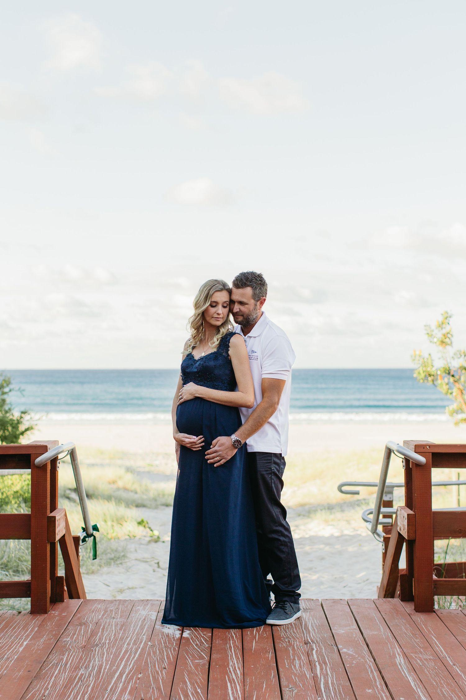 Pregnant couple embracing on a wooden beach deck, woman in navy blue maternity gown, ocean view behind them.