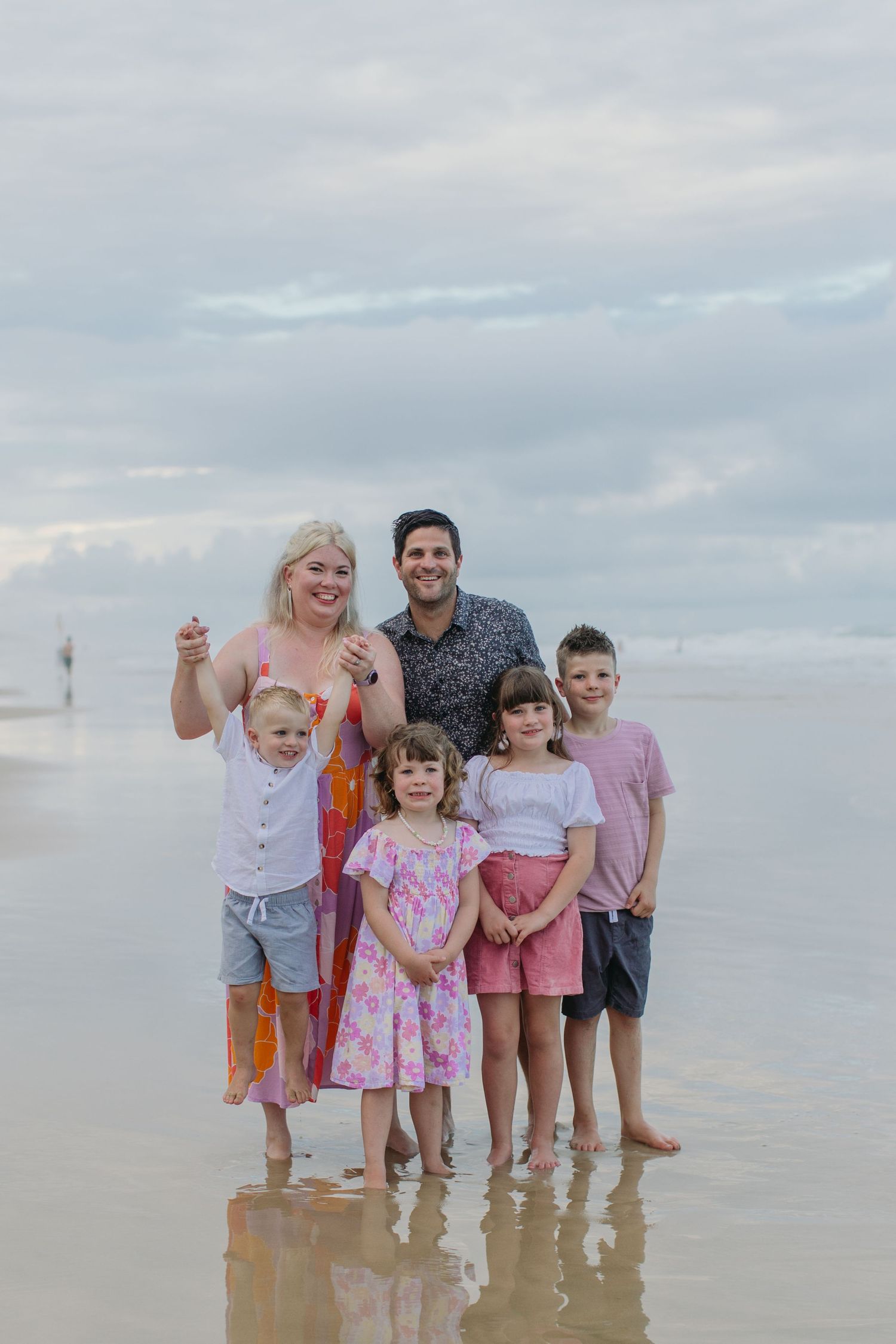 Family of six posing on a reflective beach with cloudy skies, two parents and four children smiling together.