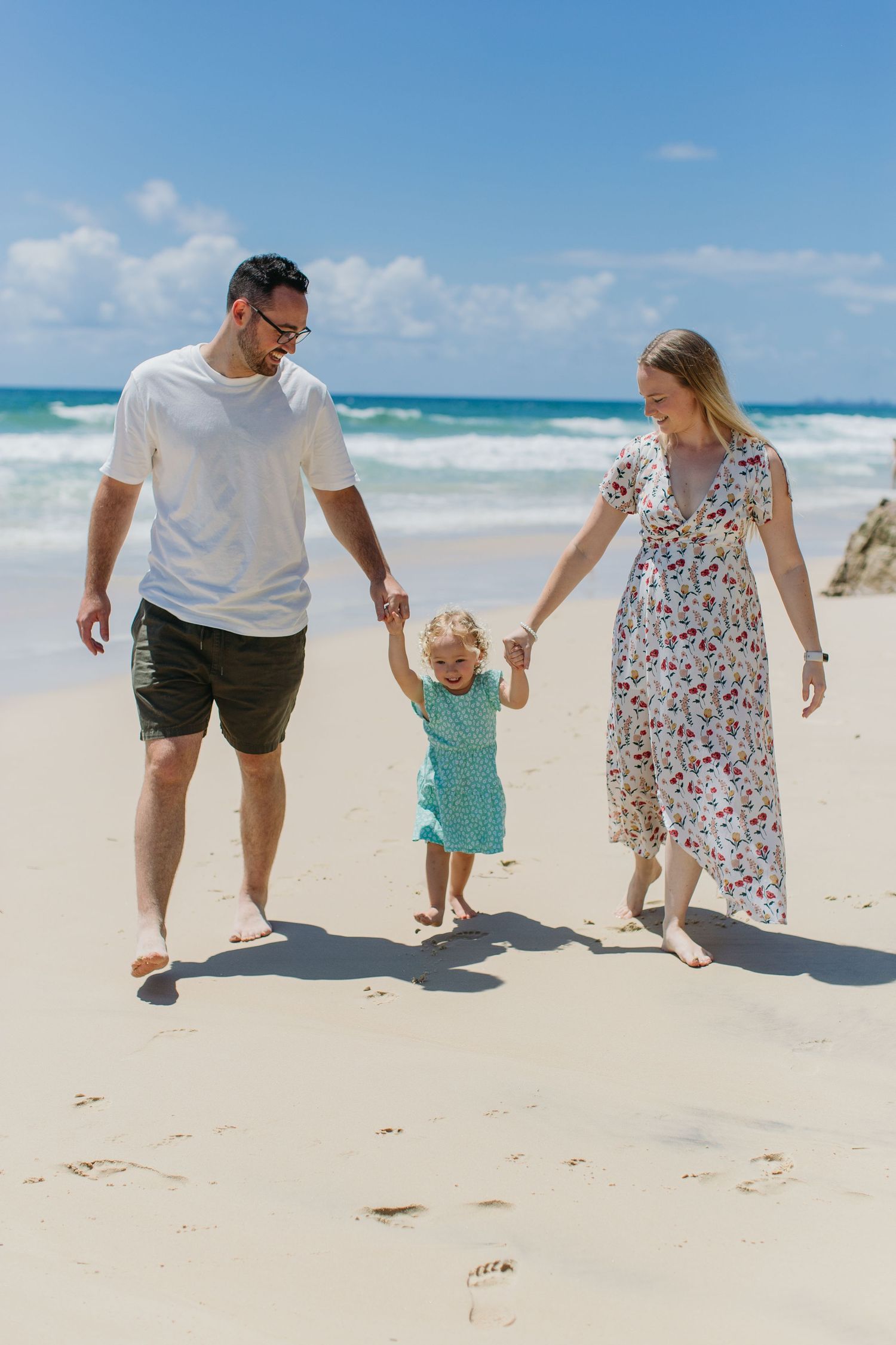 Family of three walking hand in hand on a sunny beach, parents holding toddler girl in teal dress.