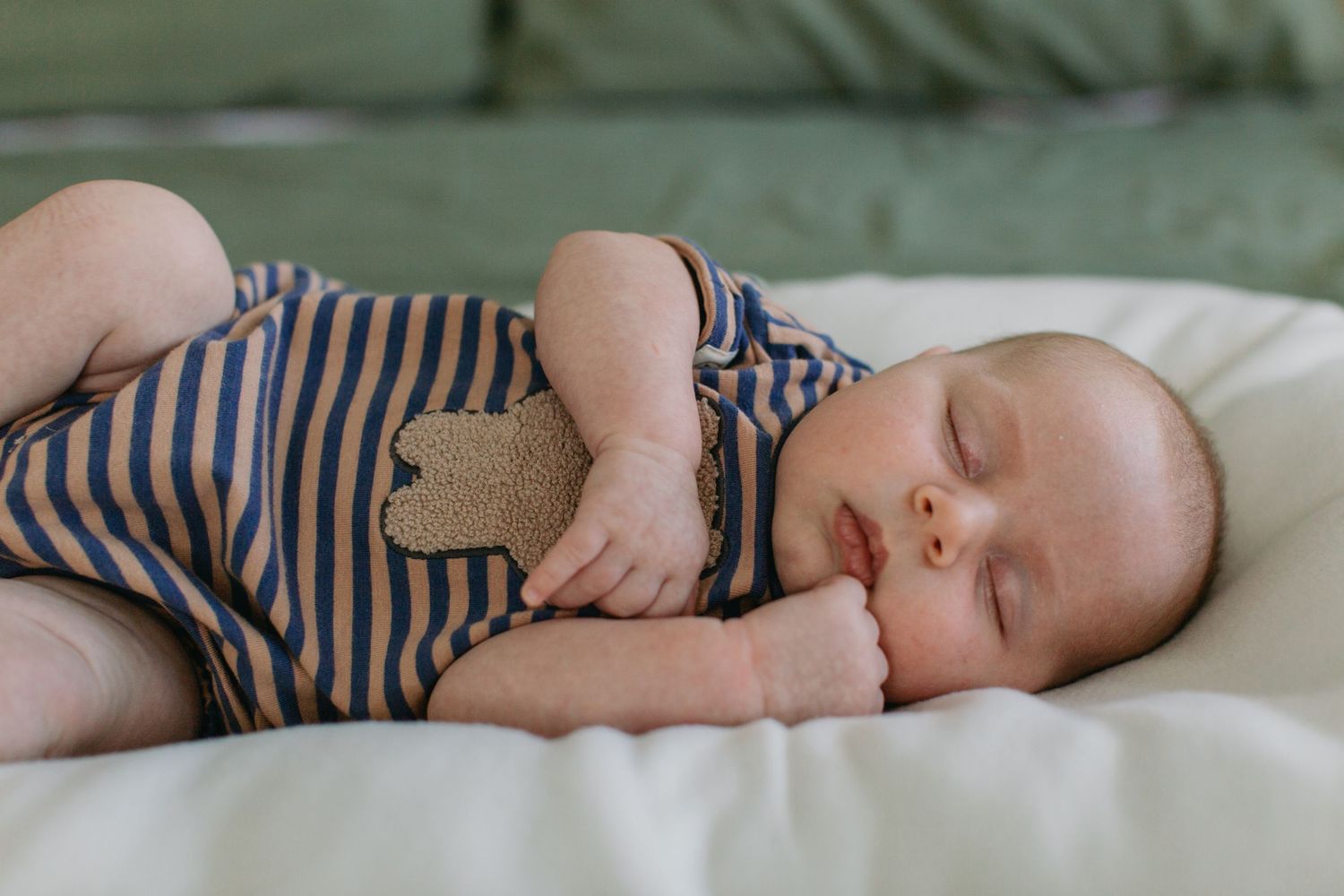 Peaceful newborn baby sleeping on a cream pillow wearing a blue striped romper, holding a small stuffed toy.