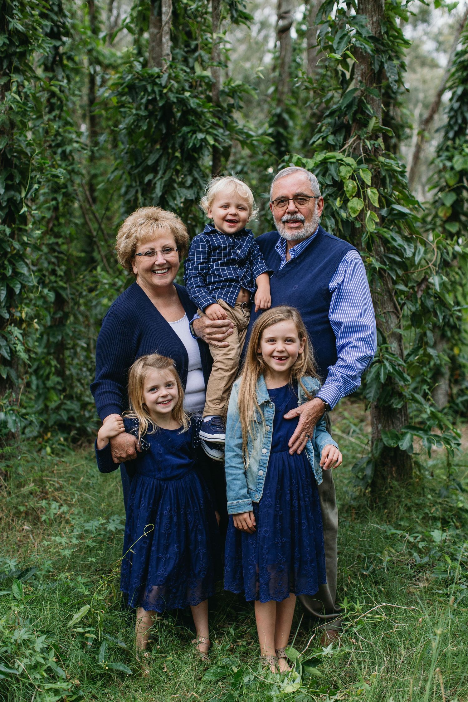 Grandparents posing outdoors with three young grandchildren in matching navy outfits among lush green trees.