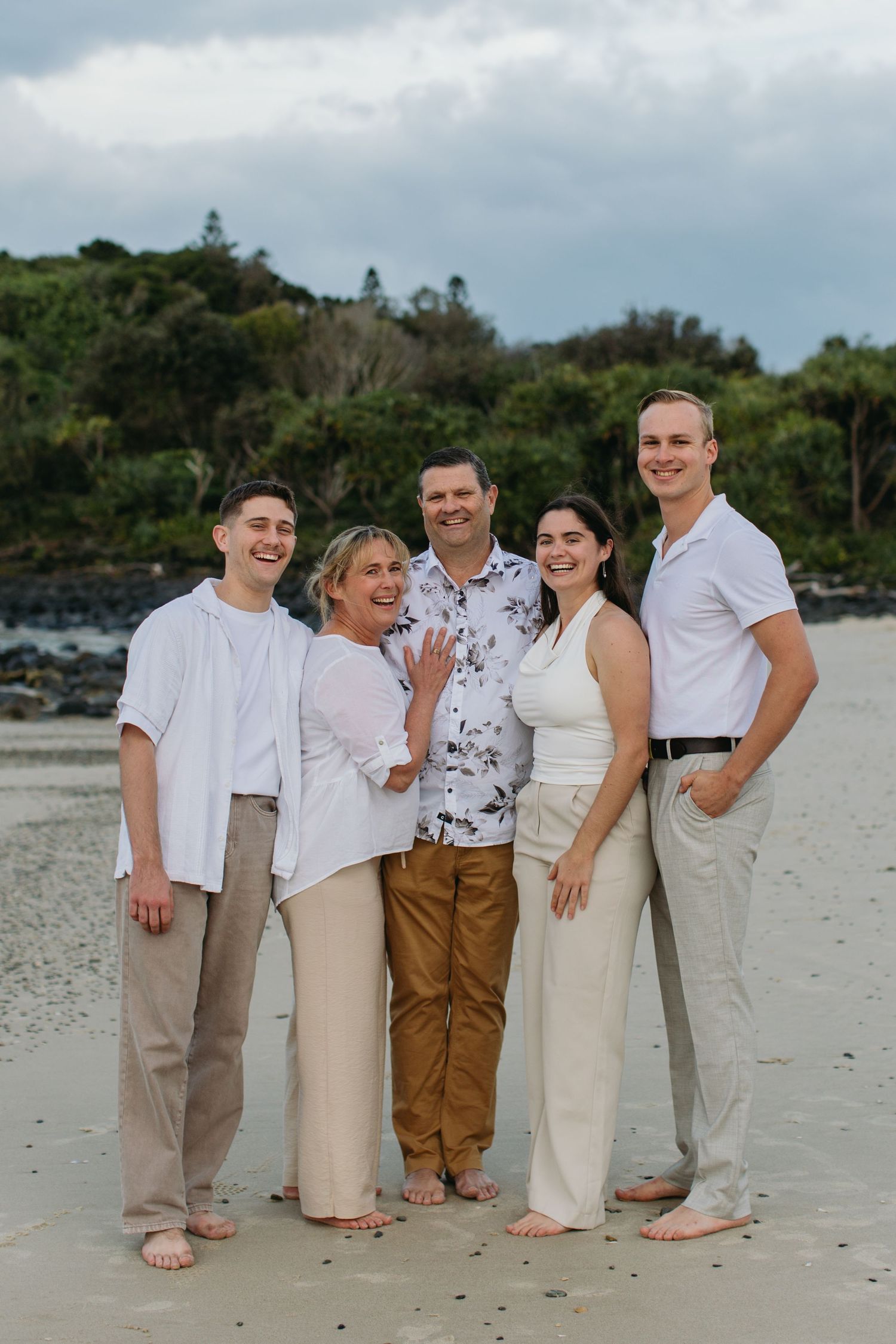 Family of five posing together on a sandy beach, smiling, dressed in white and neutral tones with lush greenery behind.