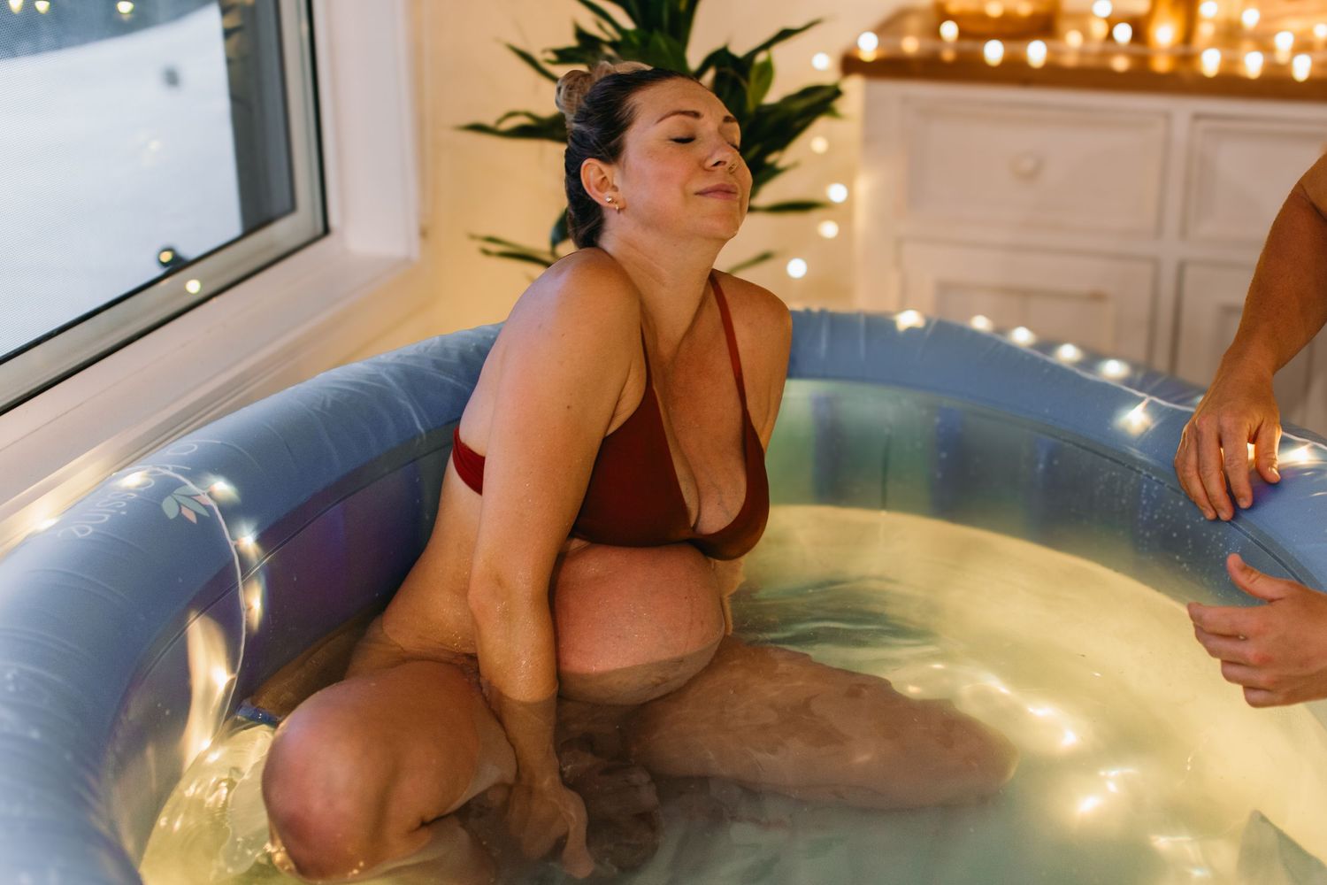 Woman in red bikini top relaxing in an inflatable birthing pool surrounded by fairy lights during labor.