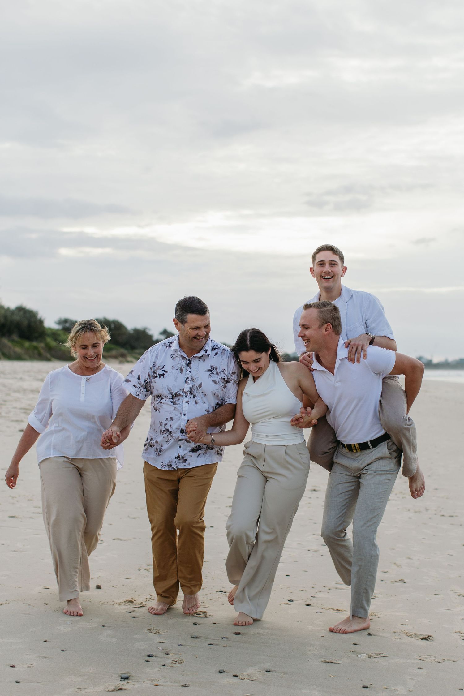 Five adults laughing and walking barefoot on a sandy beach, with a man giving a piggyback ride under a cloudy sky.