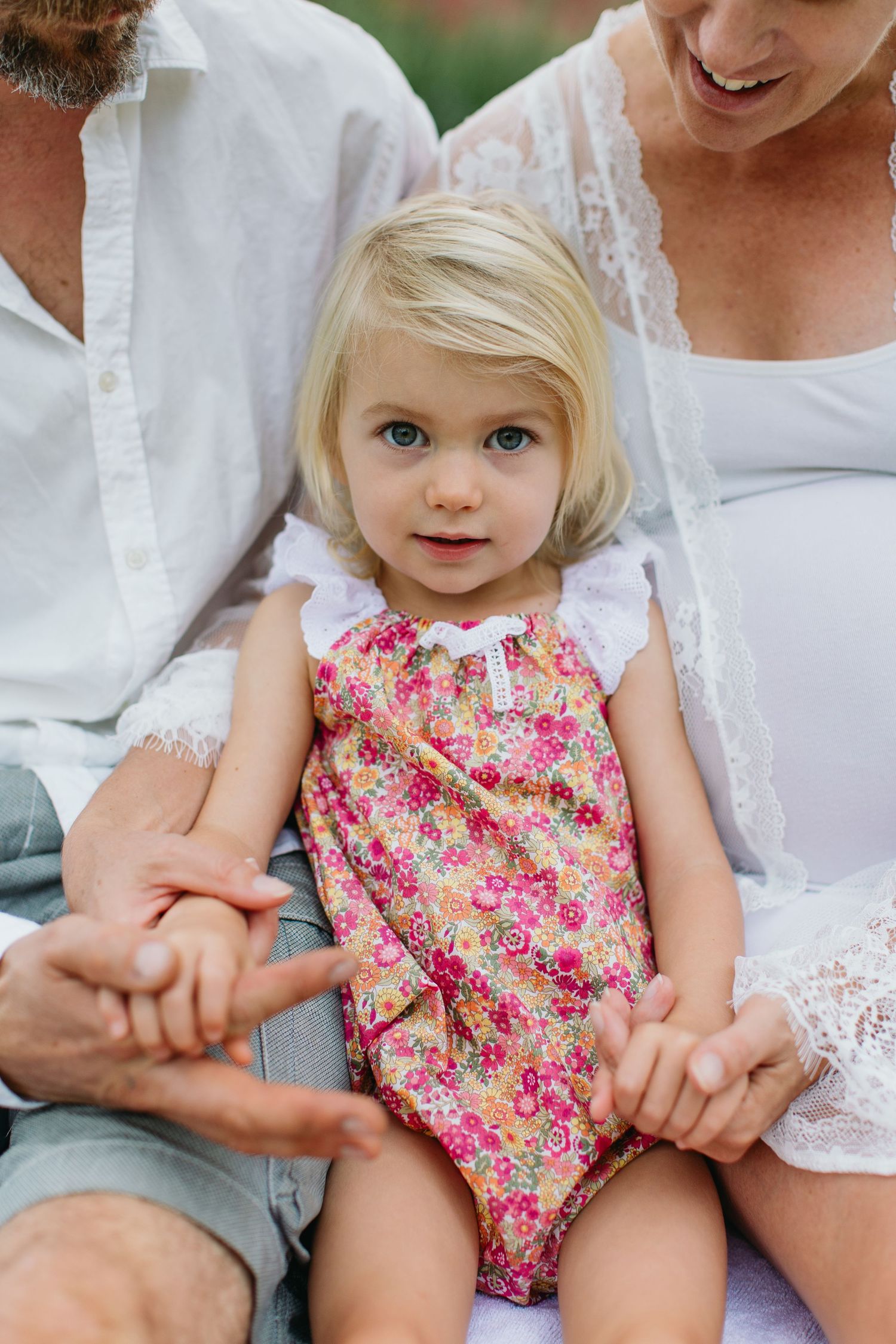 Blonde toddler girl in floral romper sitting with parents outdoors during a family maternity photo session.
