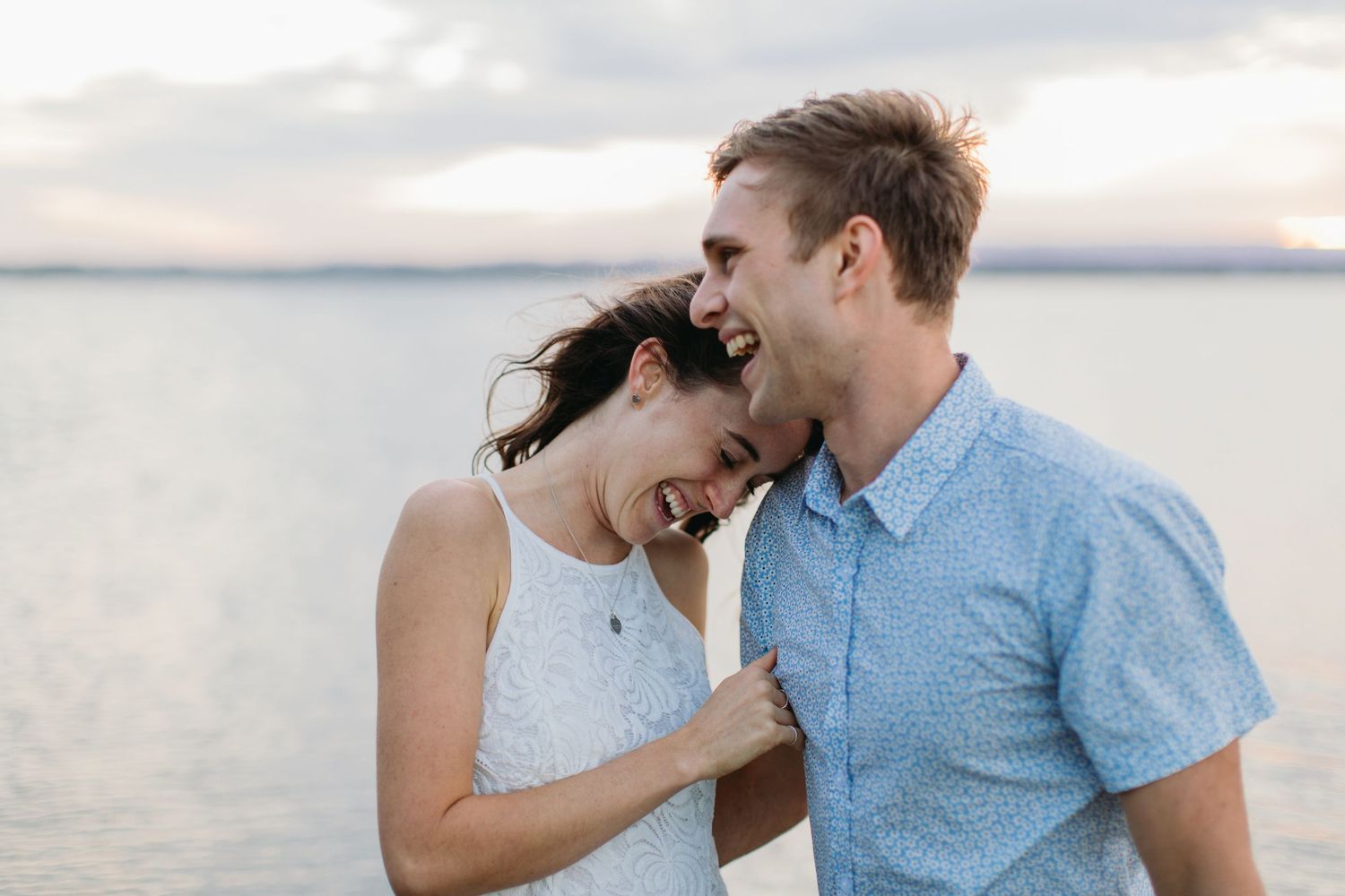 Happy couple laughing together on a beach at sunset, sharing a joyful romantic moment by the water.