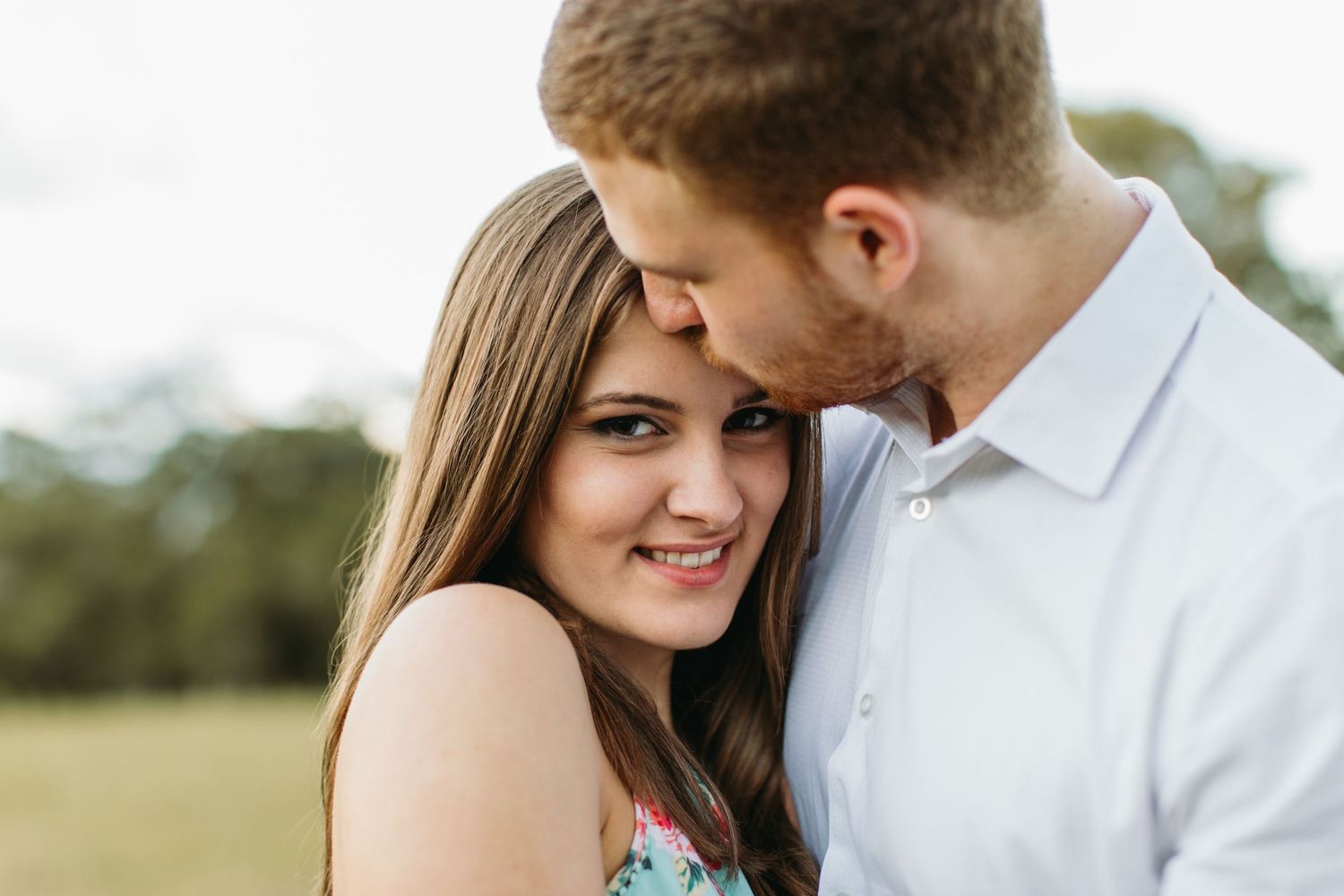 Romantic couple sharing a tender moment outdoors, man kissing woman's head while she smiles in a green field.