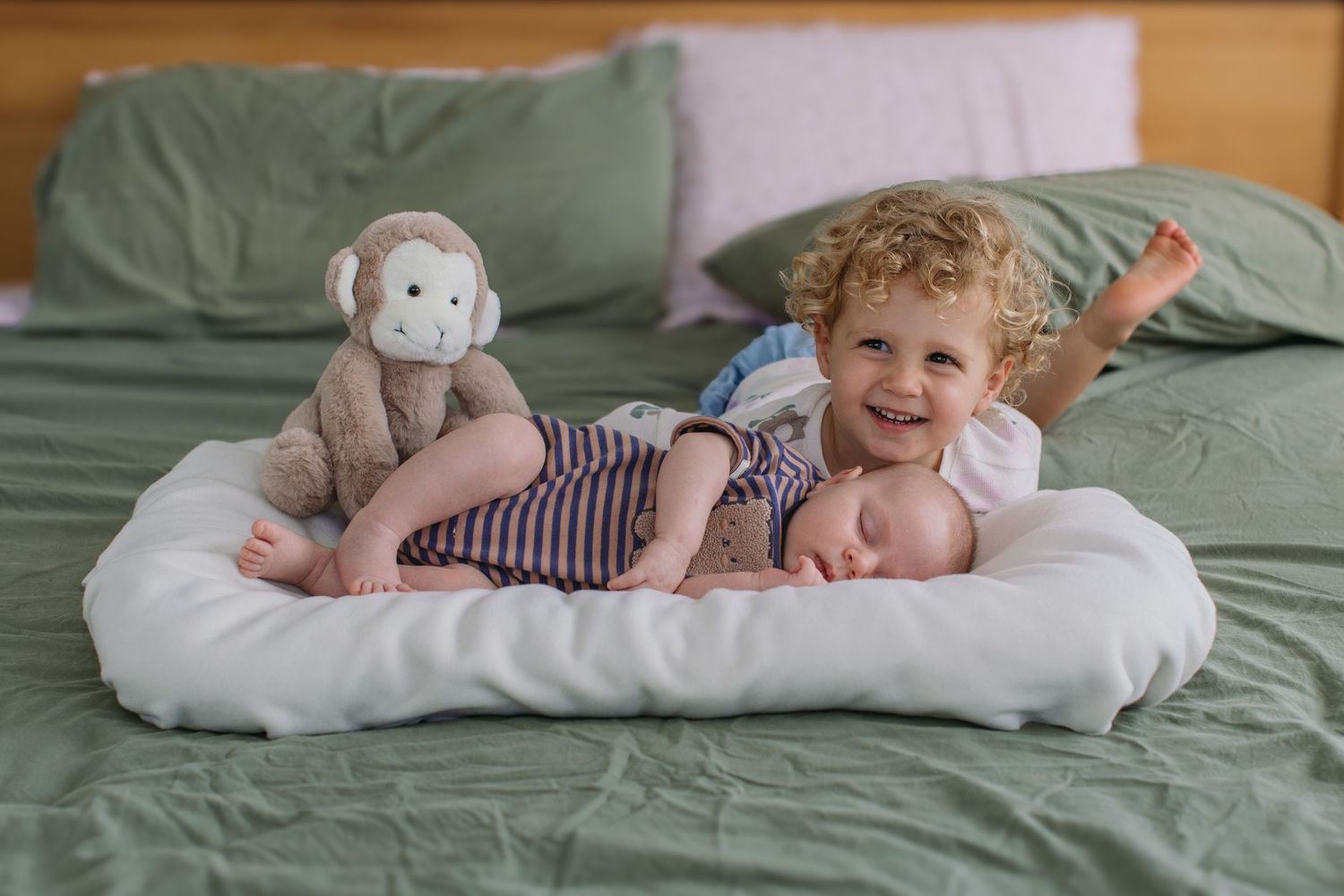 Toddler with curly hair smiling beside sleeping newborn baby on white pillow with stuffed monkey toy on green bed.