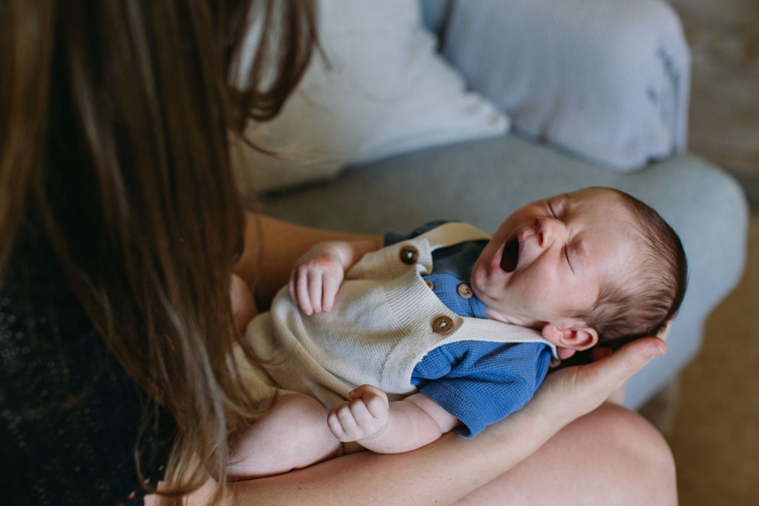 Mother holding crying newborn baby dressed in white onesie with blue accents during indoor lifestyle photoshoot.