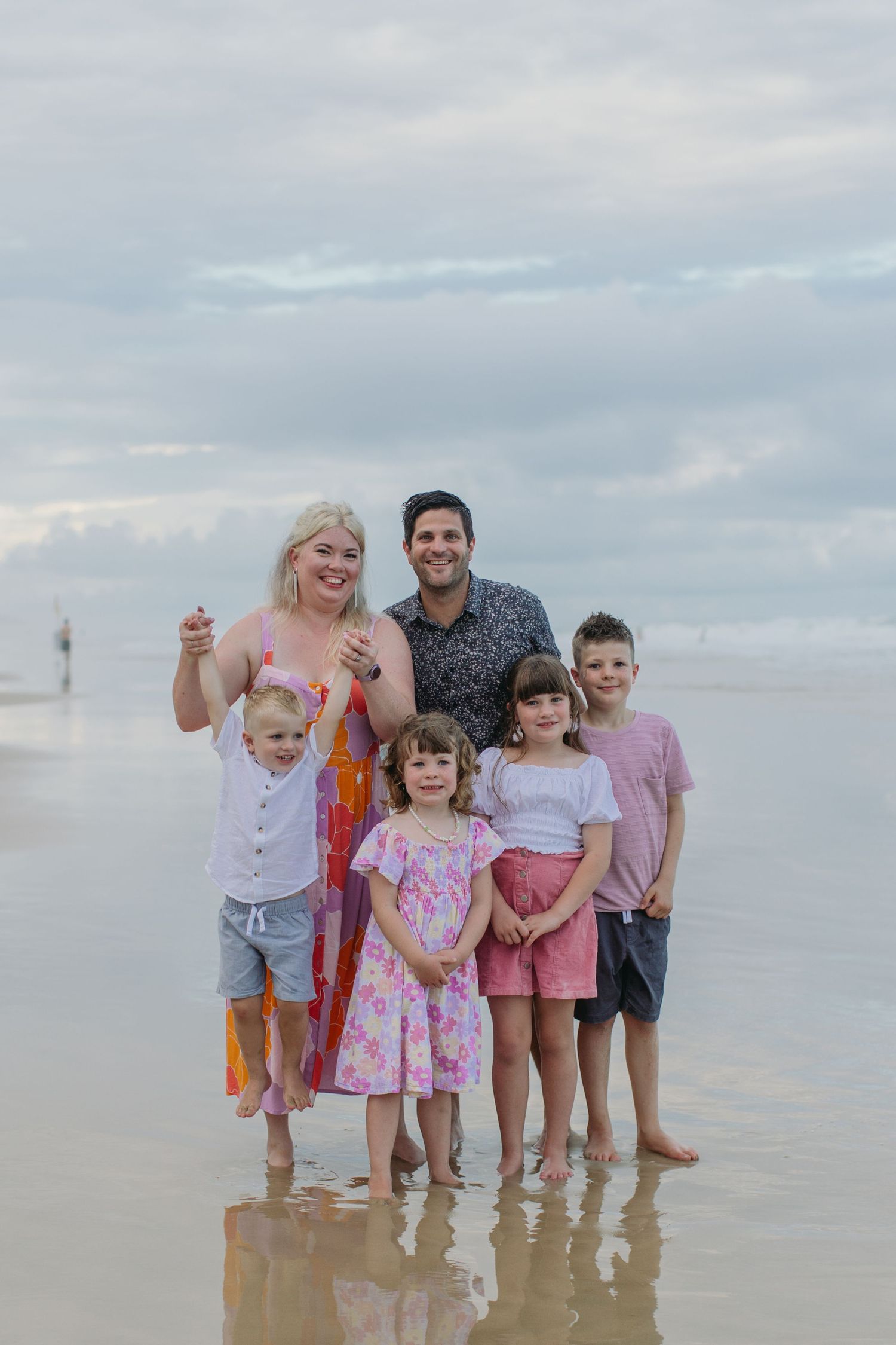 Family of six posing on a beach shoreline, two adults and four children smiling, overcast sky in the background.
