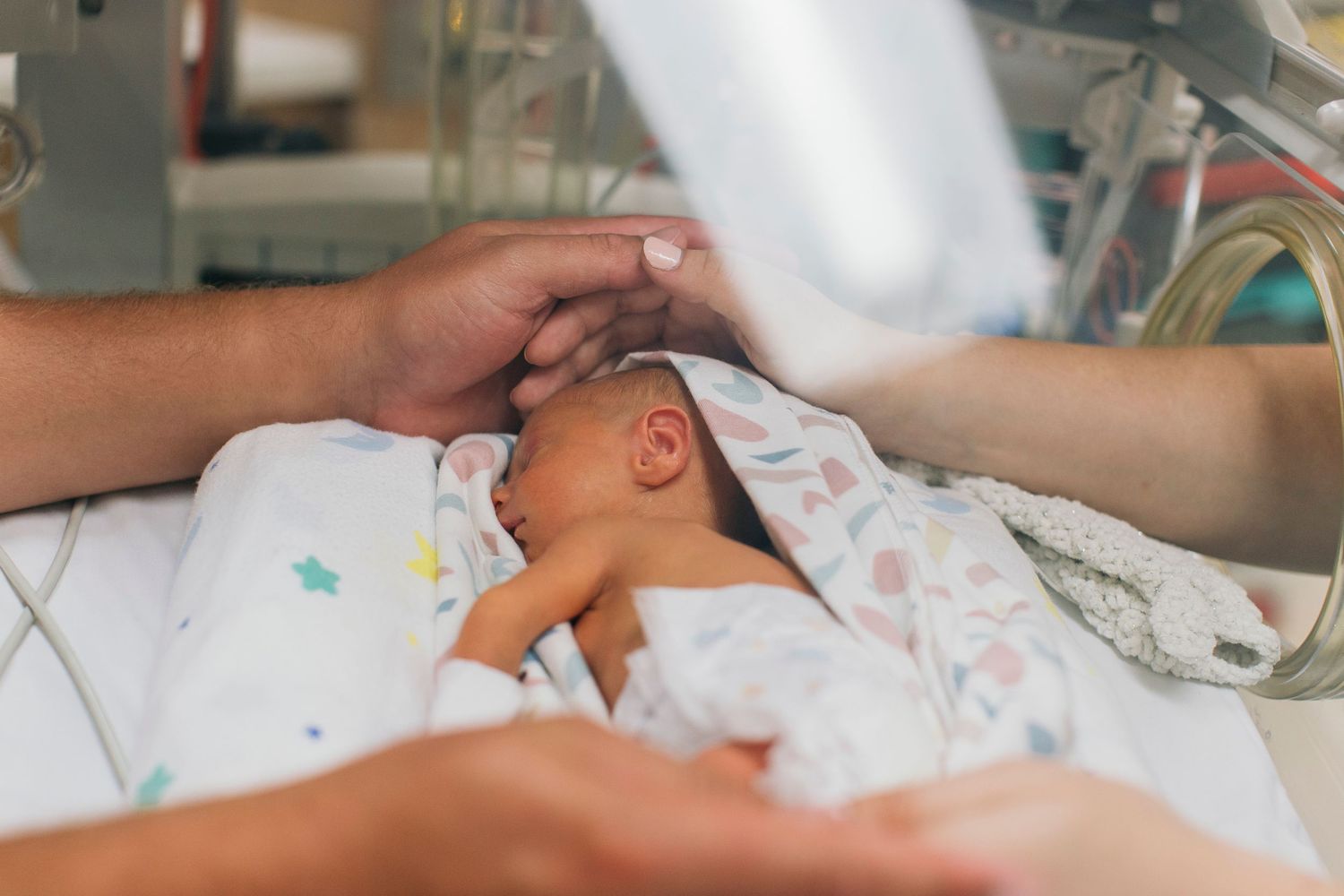 Premature newborn baby resting in hospital incubator with parents gently touching their infant.
