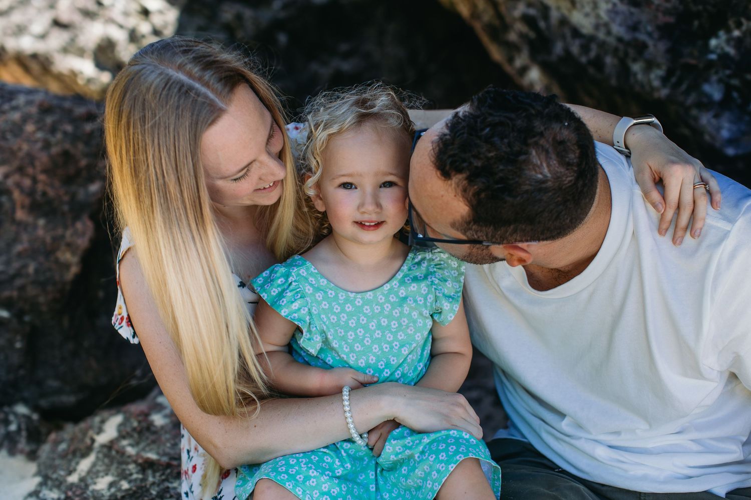 Family portrait with parents hugging blonde toddler girl near rocky beach.