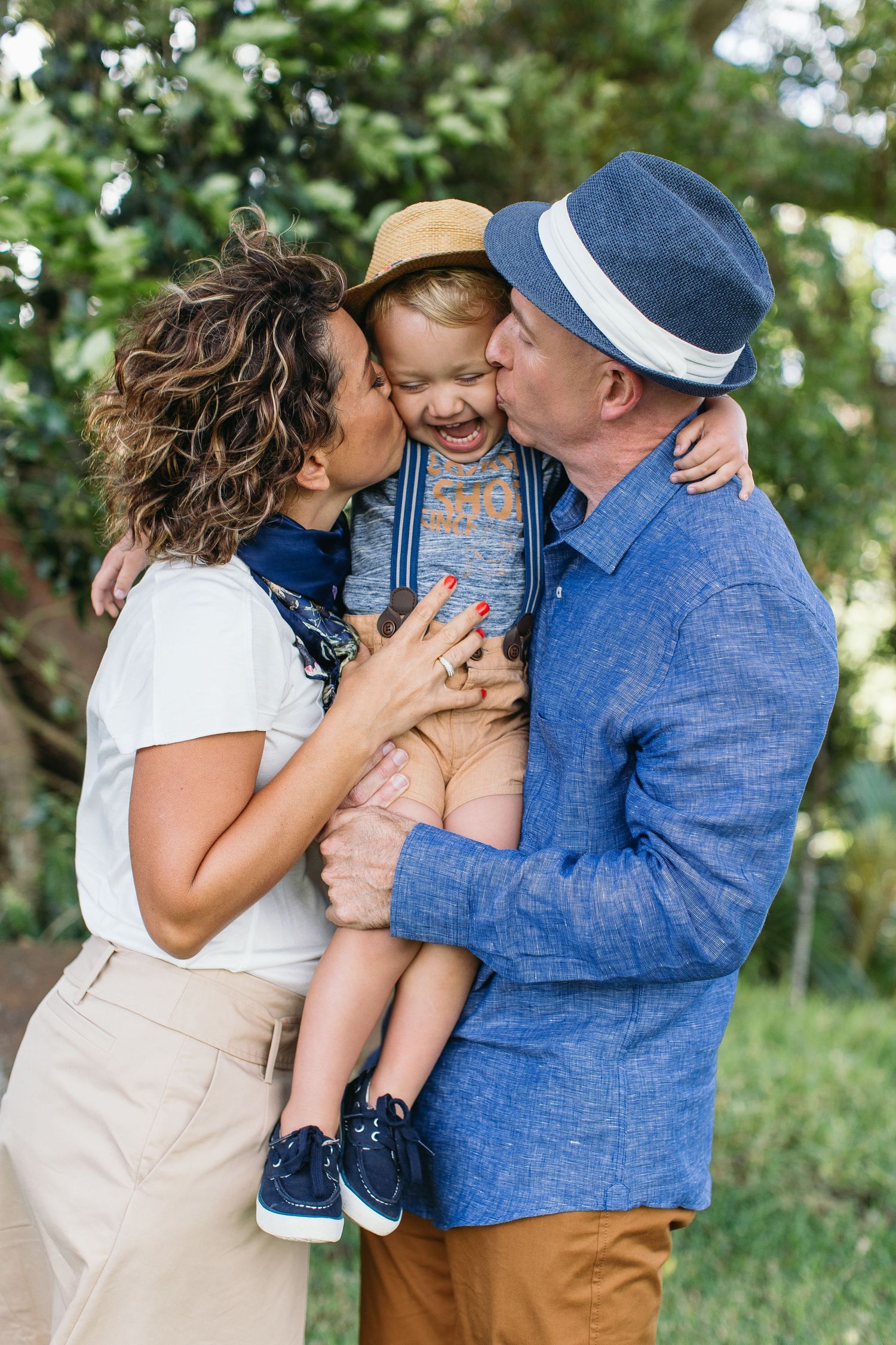 Happy family with parents kissing their laughing toddler boy outdoors in a lush green park setting.