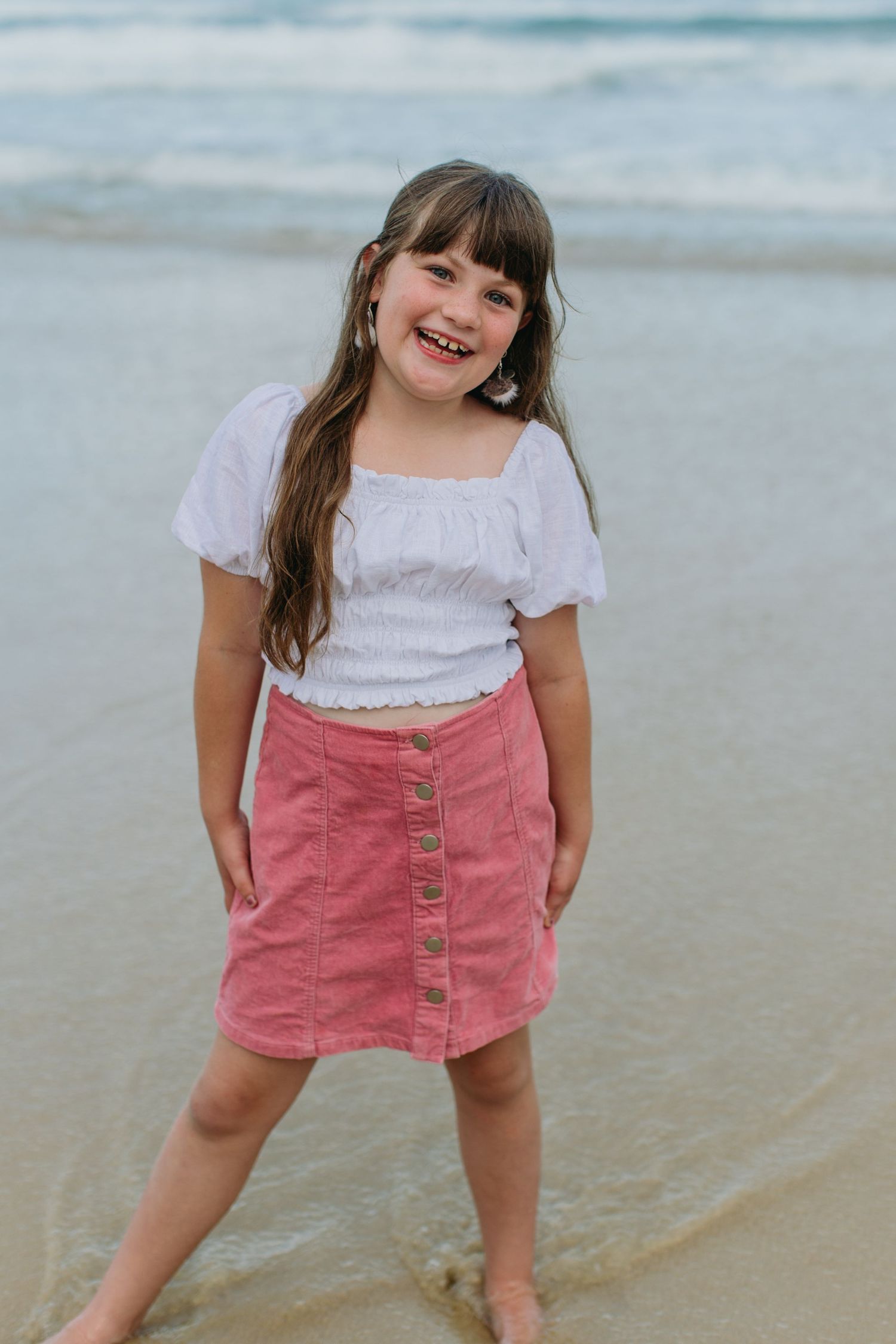 A happy young girl in a white top and pink button skirt smiles at the camera while standing at the beach.