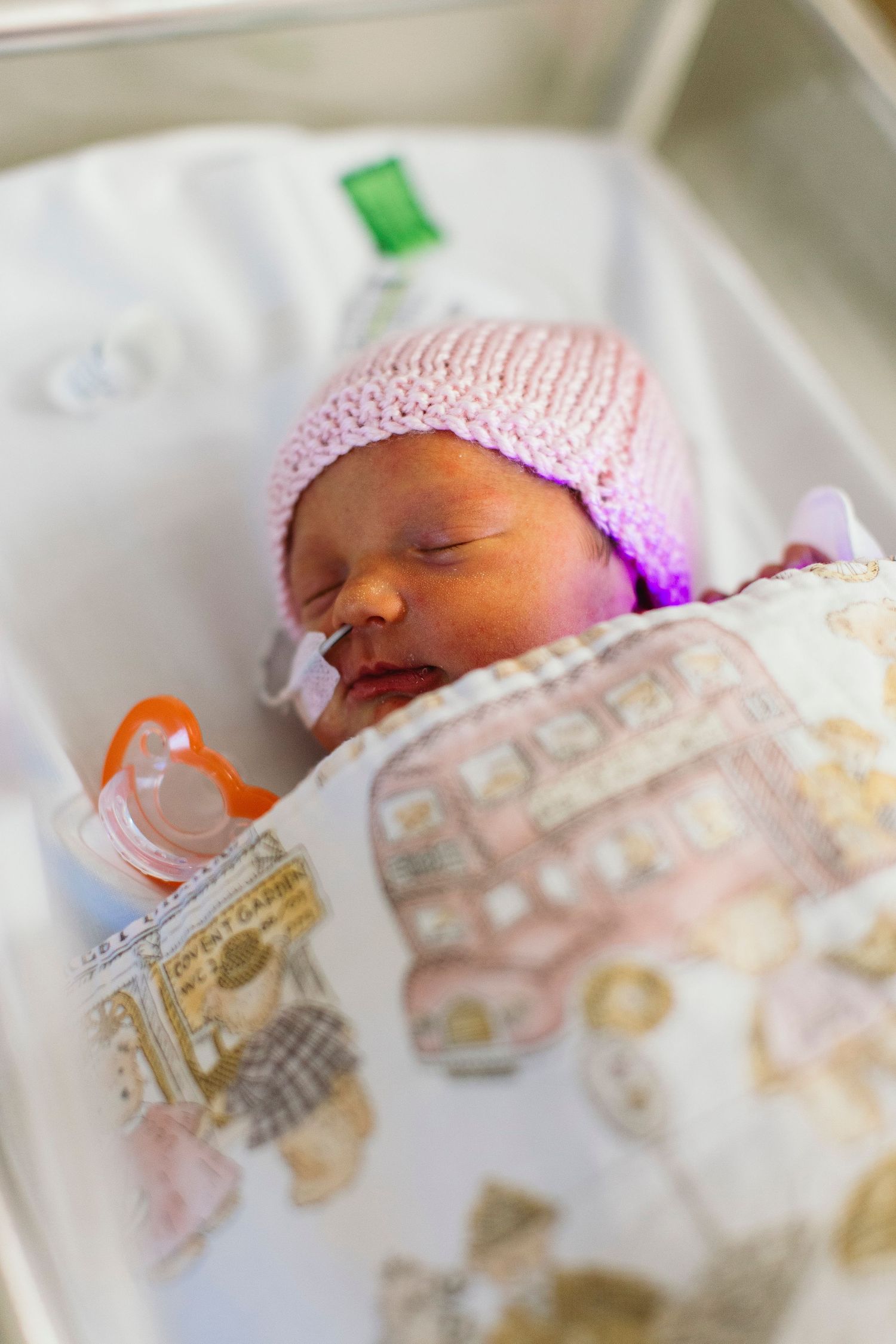Newborn baby sleeping in hospital bassinet wearing pink knit hat, wrapped in patterned blanket with pacifier nearby.