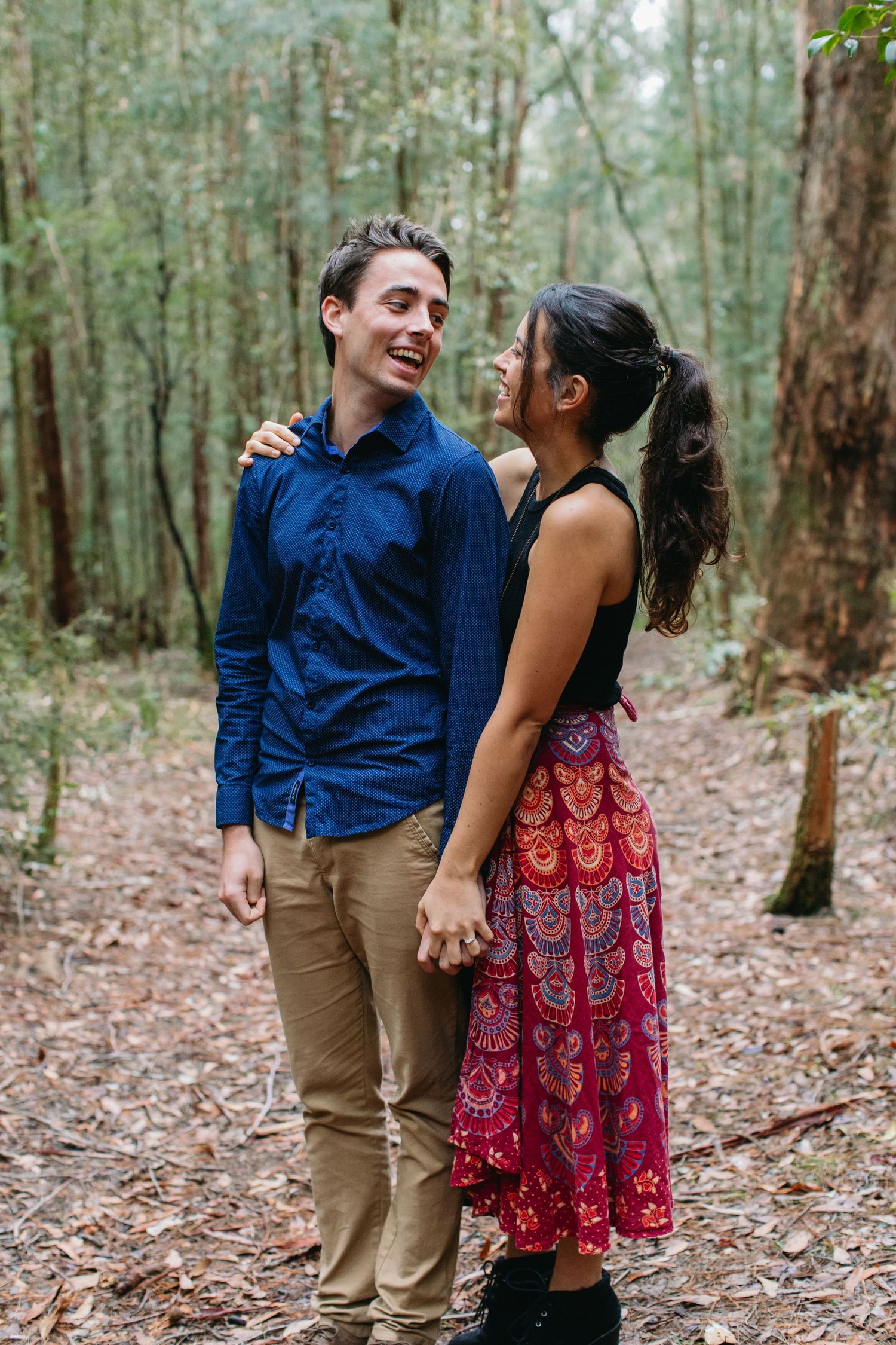 Couple laughing and holding hands in a forest, man in blue shirt and woman in colorful red skirt.