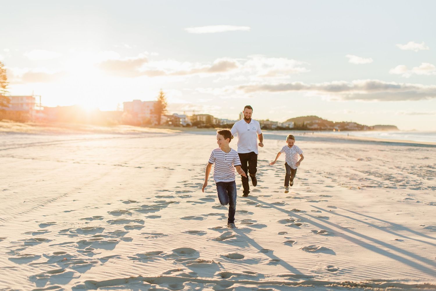 A father and two children run joyfully on a sandy beach at golden sunset with ocean views in the background.