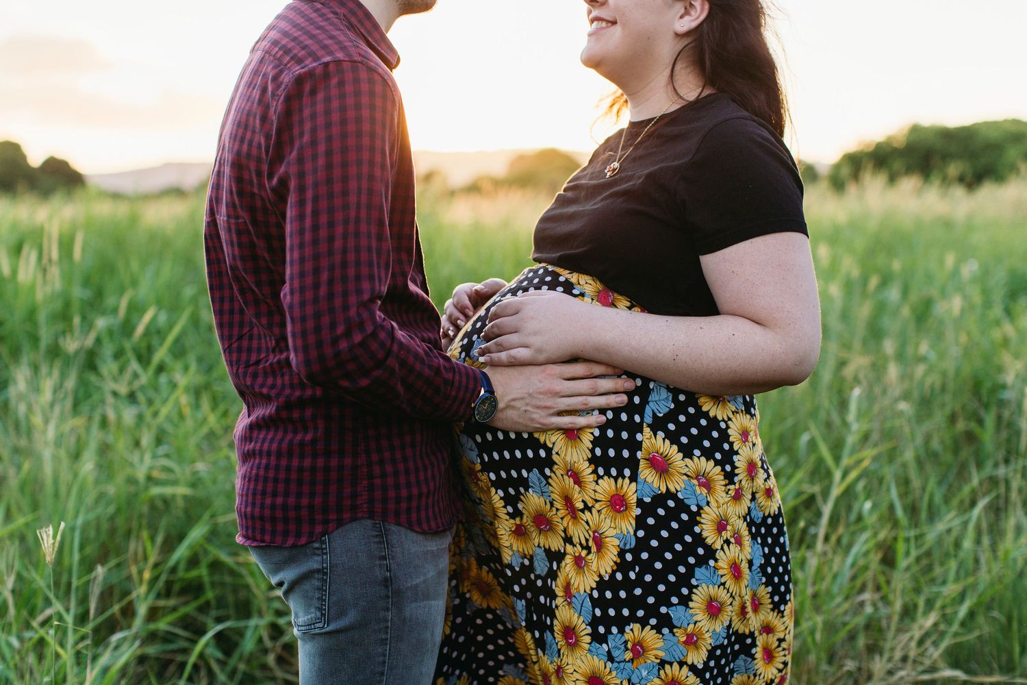 Pregnant couple holding baby bump in sunflower skirt at golden hour in a green field.