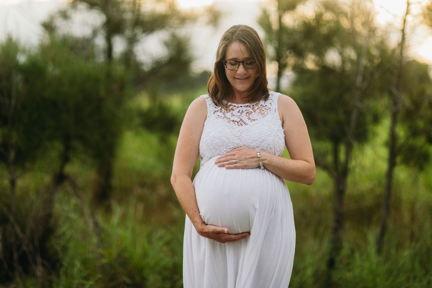 Pregnant woman in white lace dress cradling baby bump outdoors in green nature setting during golden hour.