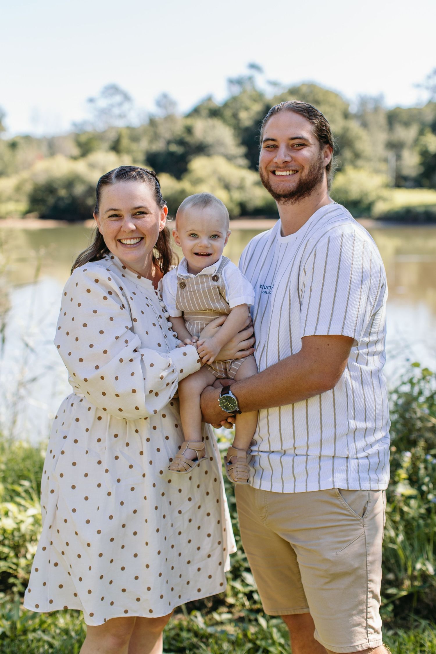 Happy young family with baby posing outdoors near a lake surrounded by lush greenery on a sunny day.